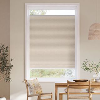 A minimalist dining space with a large window framed in white. A beige pleated blind covers most of the window, allowing light to filter through the lower section.