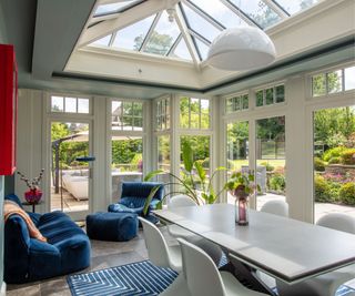 A bright conservatory with a glass roof, navy lounge chairs, a modern dining table, and white chairs. Large windows overlook a lush garden, seamlessly connecting indoor and outdoor spaces.