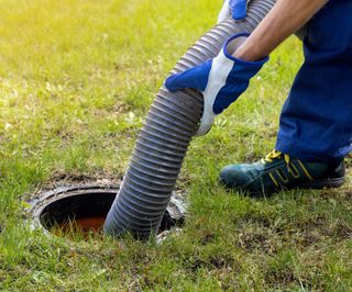 A man holding a black pipe into a hole with a septic tank inside