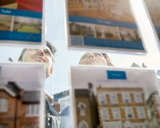 Estate agent’s window from inside with for sale details and man and woman outside looking at them
