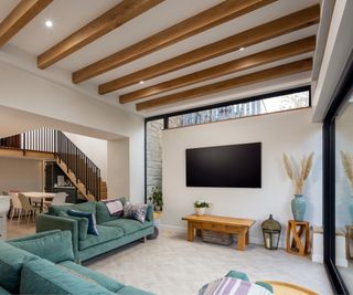inside living room extension with a modern finish, exposed timber beams, glazing above tv wall, dropped arch separating living room from kitchen and dining area, green sofas, wooden floor