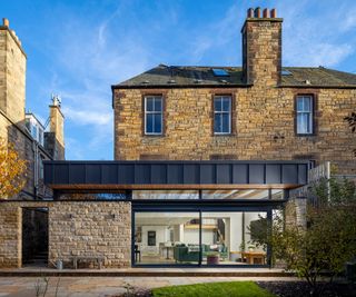 exterior of large stone house with modern extension to rear with clad flat roof, large sliding patio aluminium doors, stone wall to side and patio area to front