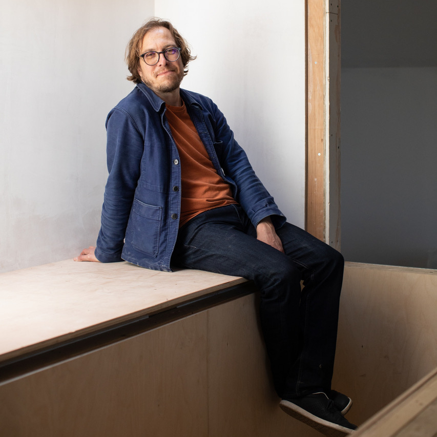 male with glasses, facial hair, wearing denim jacket, black trousers and orange top sat on wooden cupboards