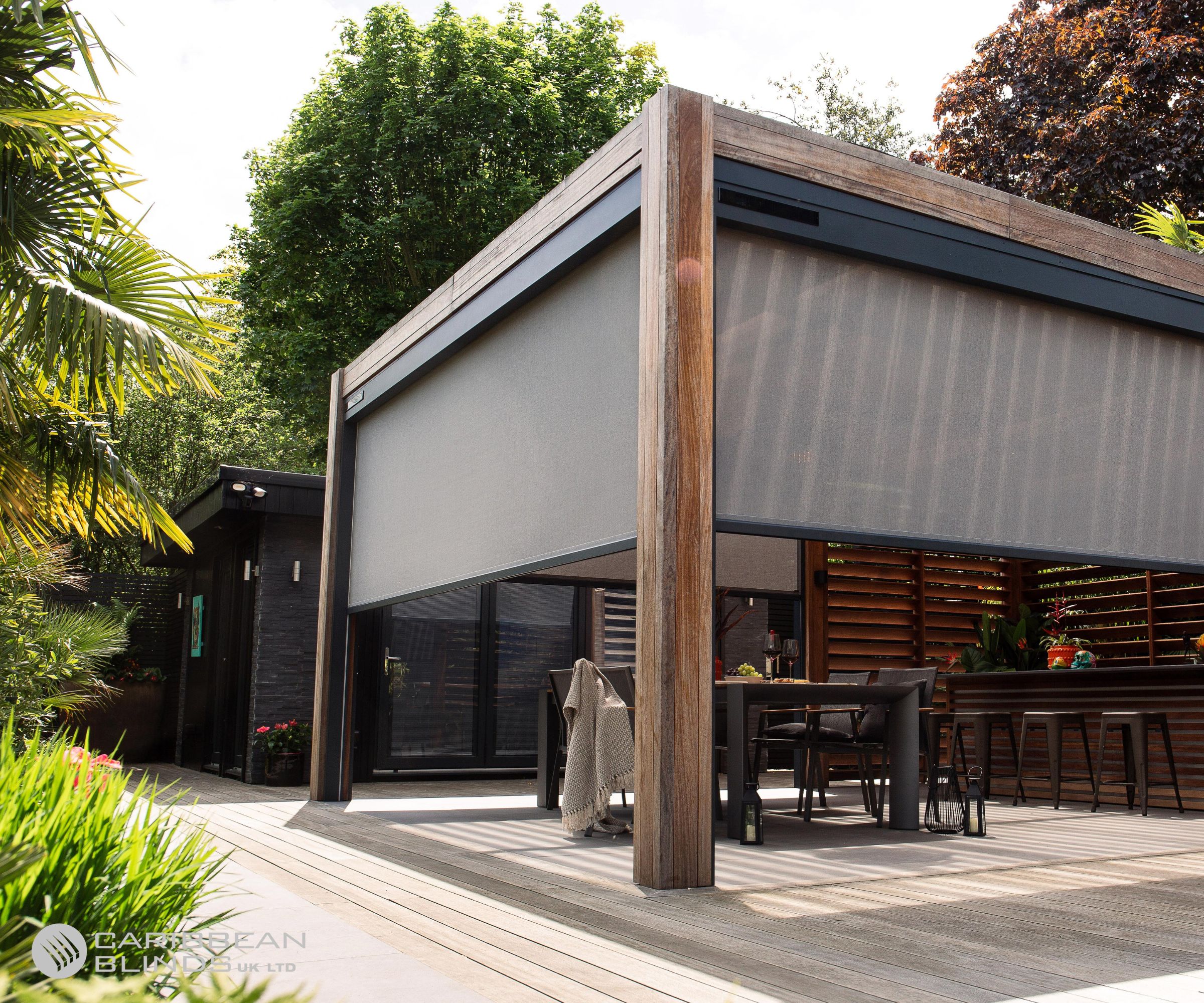 A sleek, contemporary pergola with a slatted wooden frame and grey retractable blinds shades a stylish outdoor dining area. Black bar stools line a counter, and lanterns decorate the light-toned decking.
