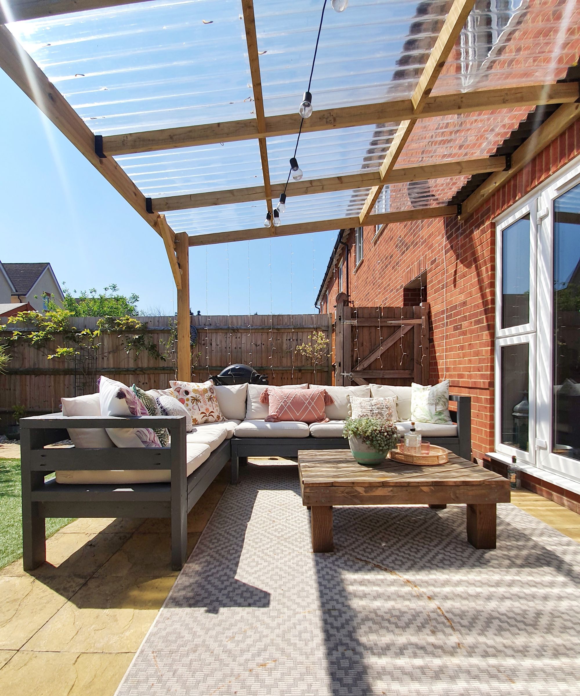 A wooden pergola with a clear corrugated plastic roof shelters an L-shaped grey outdoor sofa. Cushions in soft tones and florals add comfort. A rustic wooden table and outdoor rug complete the setting.