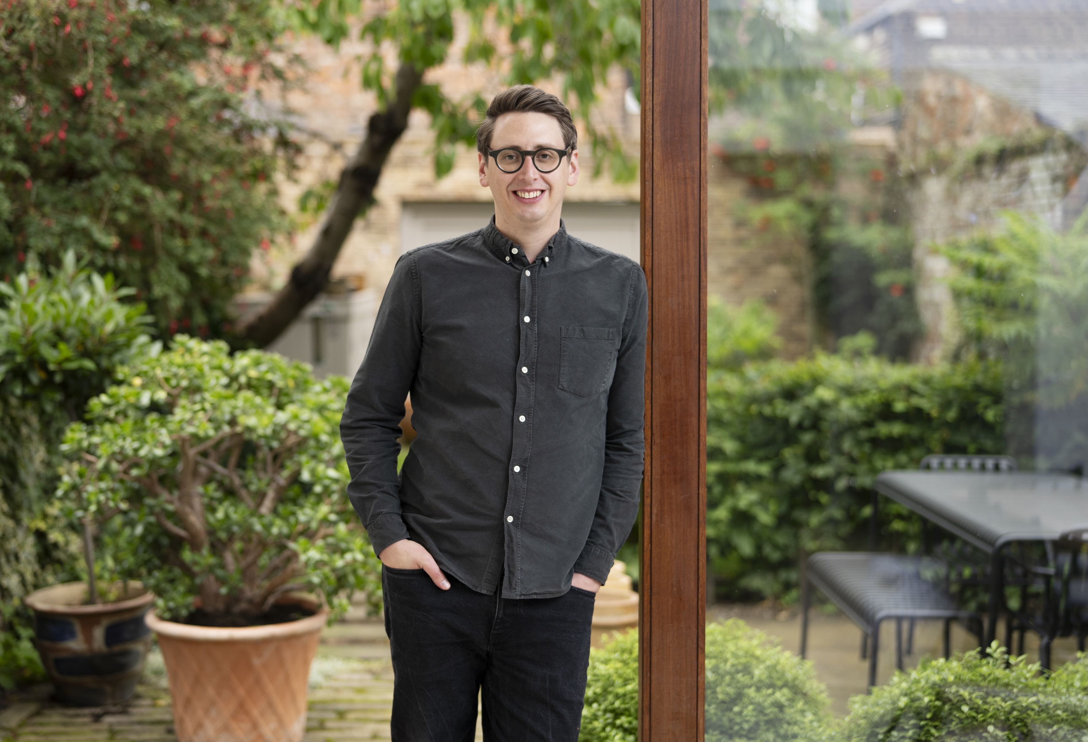 A man standing beside a wood-framed window with potted plants behind him