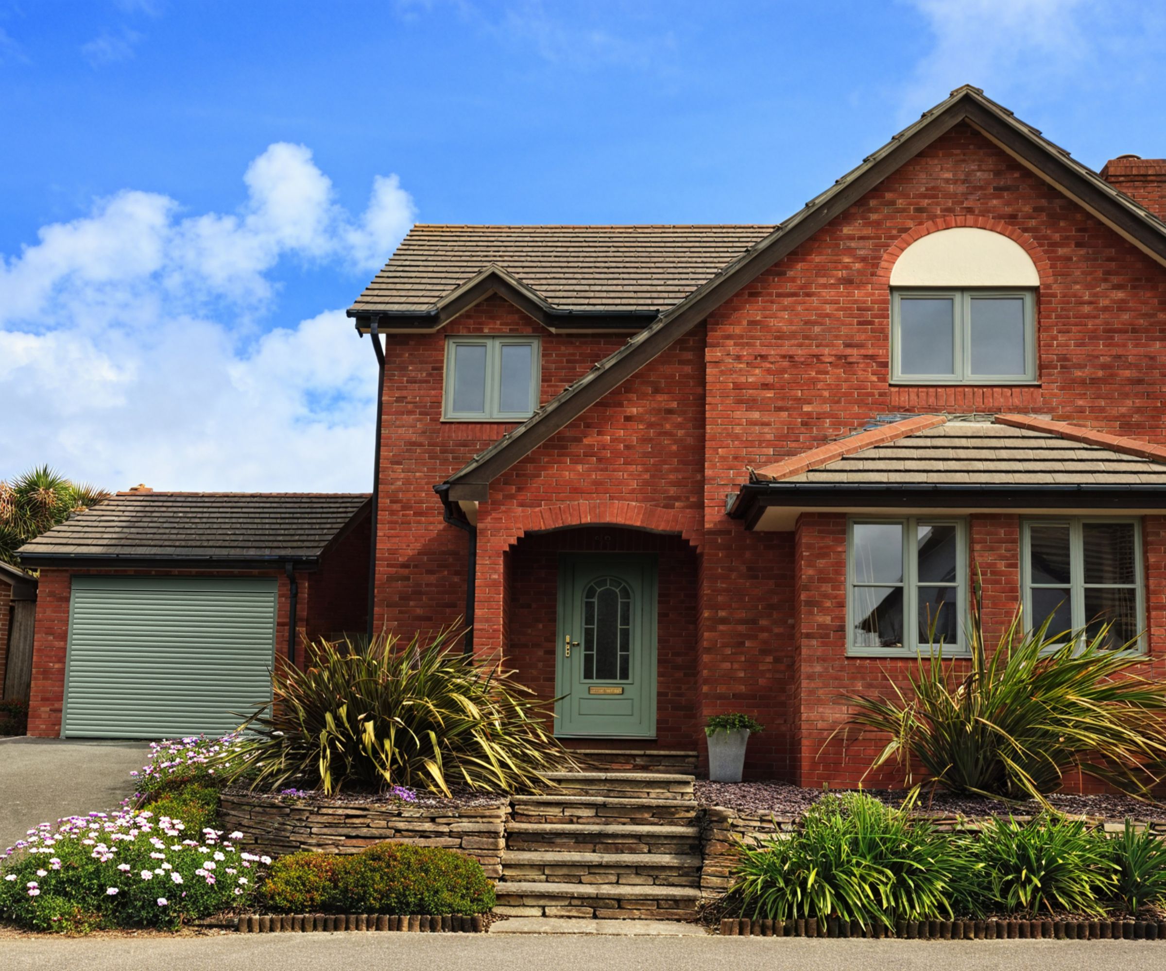 A red-brick detached house with sage green window frames and a matching roller garage door. Neatly landscaped front garden with flowering plants and stone steps leading up to a covered front door.