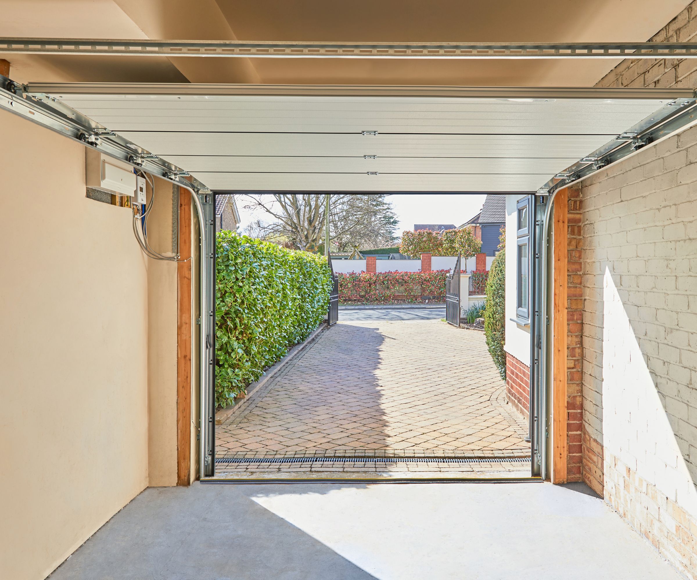 A bright garage interior with smooth concrete flooring and an open sectional garage door, revealing a paved driveway lined with green hedges and a view of neighbouring houses under a blue sky.