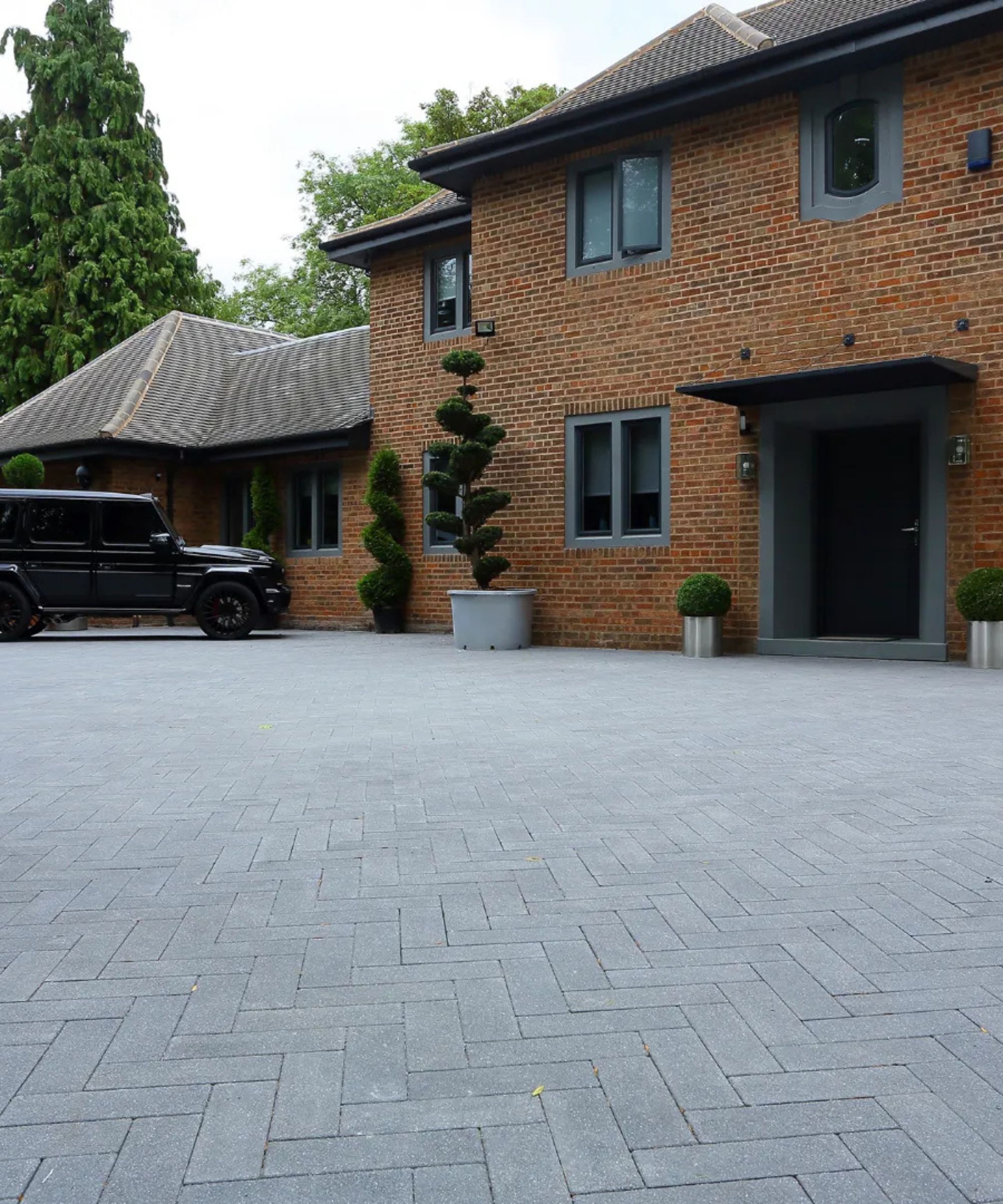 Modern brick home with dark-framed windows and a deep grey front door, complemented by a sleek grey herringbone block-paved driveway. Sculptural topiary trees in metal planters add a polished, architectural feel.