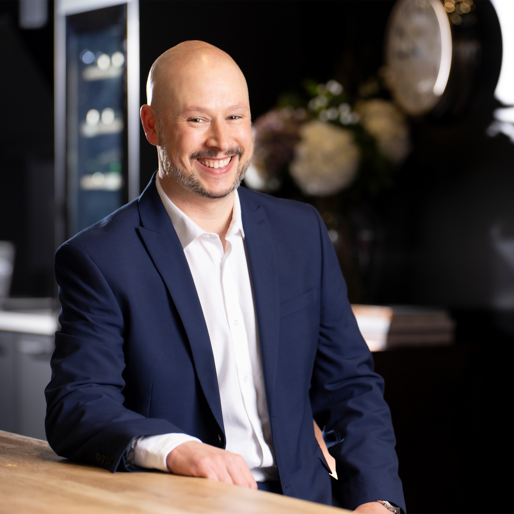 male with no hair, short beard and moustache wearing navy suit, white shirt sat at kitchen worktop