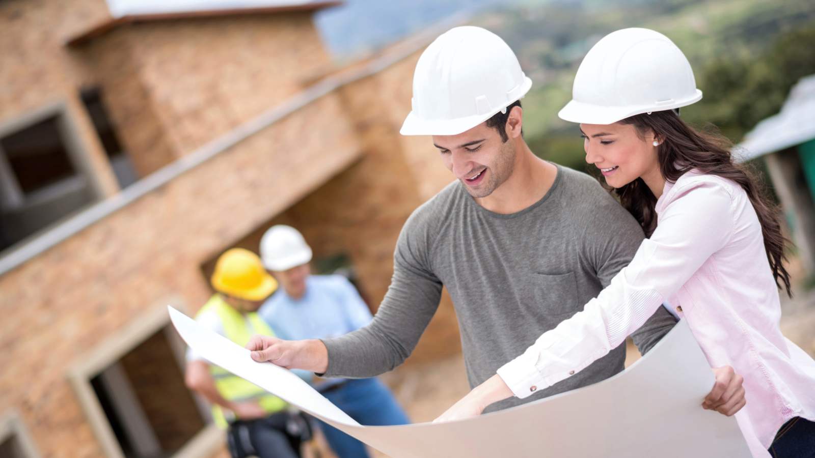 A couple in white hard hats holding architectural plans with a new build house and builders in the background