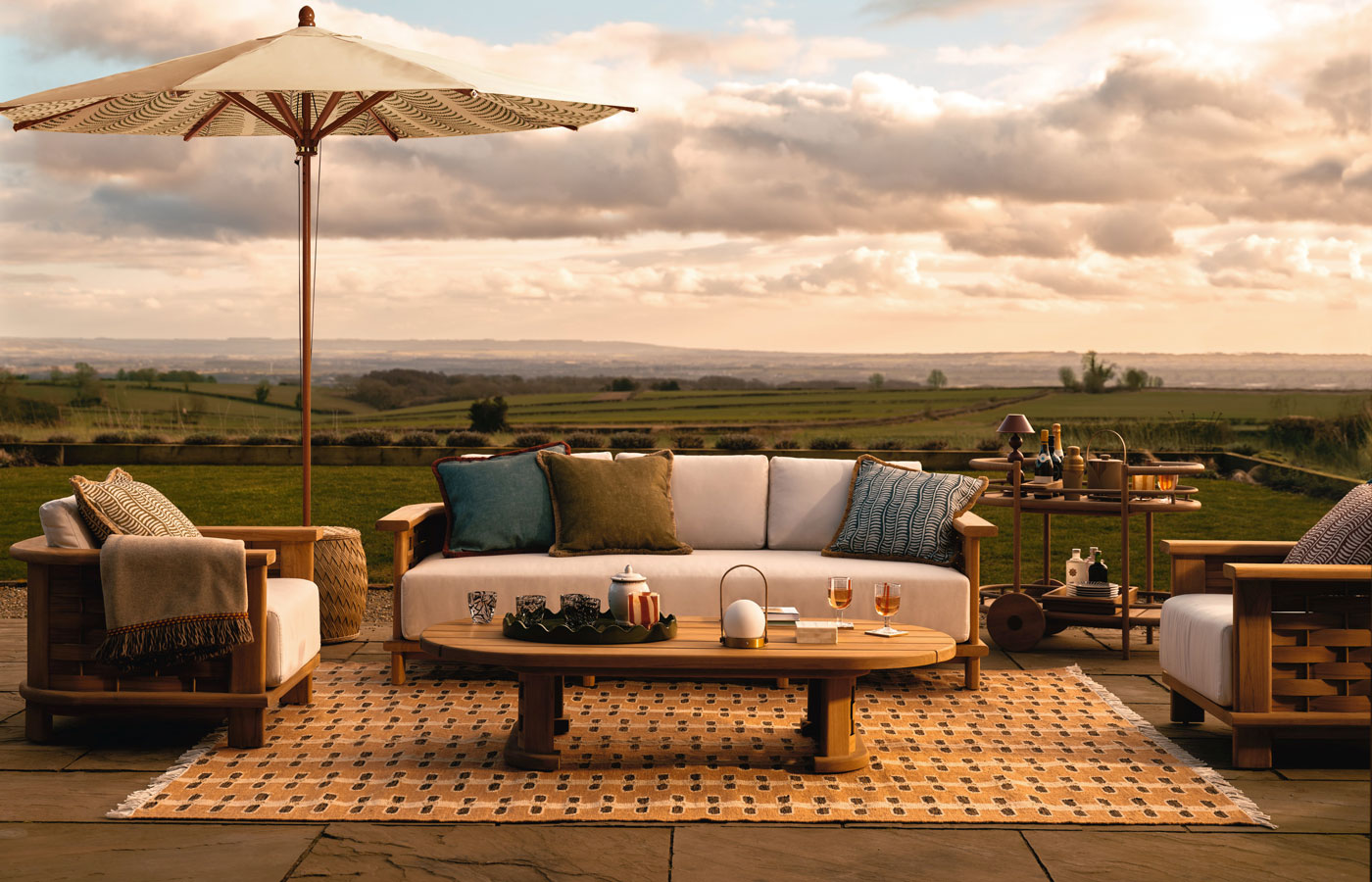 outdoor living room set-up on patio with outdoor rug, umbrella, garden sofa, garden chairs and coffee table, with a view to open countryside behind