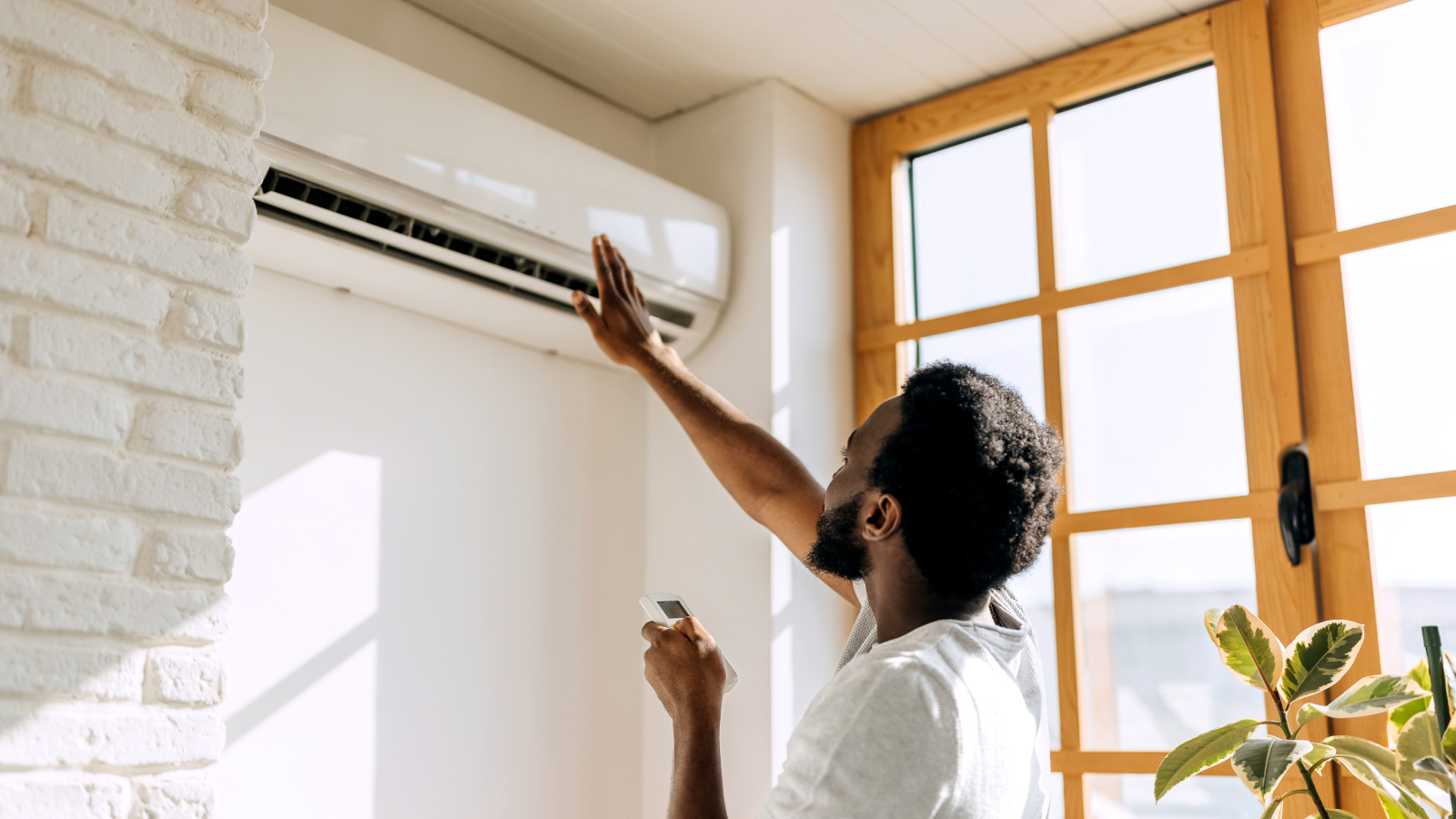 male stood facing air con unit on wall holding remote and raising hand towards unit