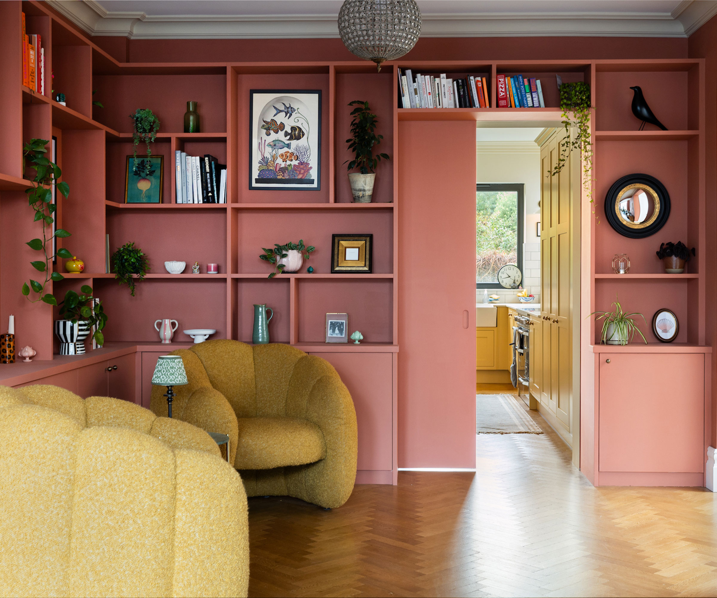 pink wall of living room shelving with herringbone wooden flooring and yellow armchairs