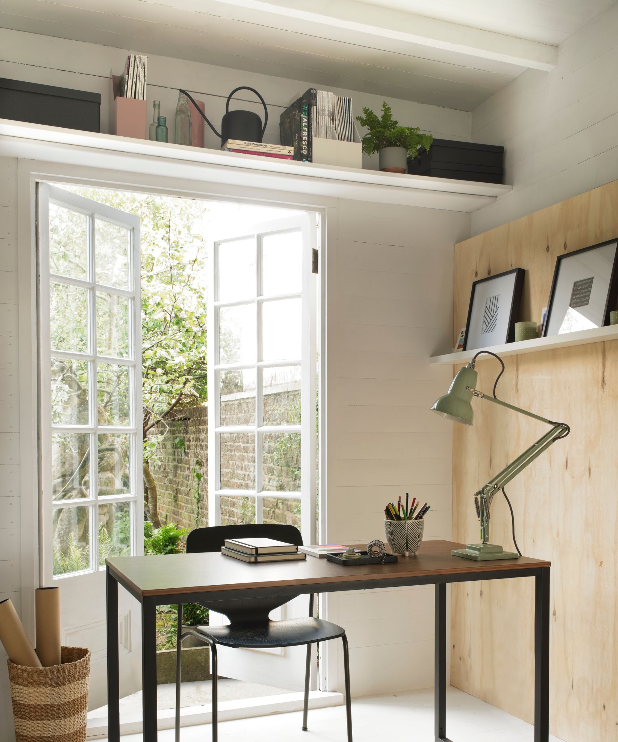 Airy garden office with French doors, minimalist desk, black chair, green desk lamp, and overhead shelves filled with books, plants, and boxes