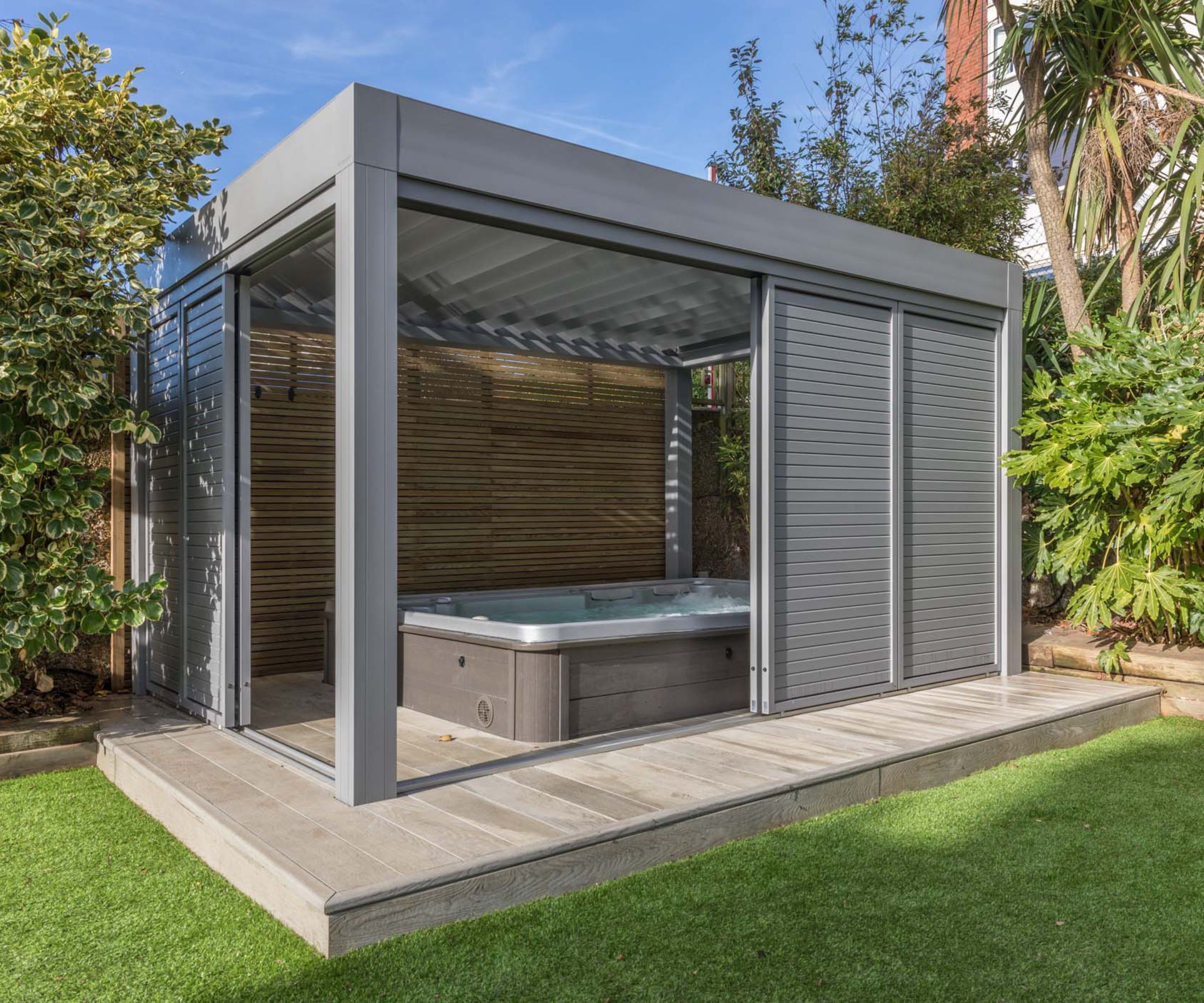 A sleek, modern garden gazebo with louvred aluminium sides and roof, housing a built-in hot tub on a raised tiled platform, surrounded by lush greenery and artificial grass.