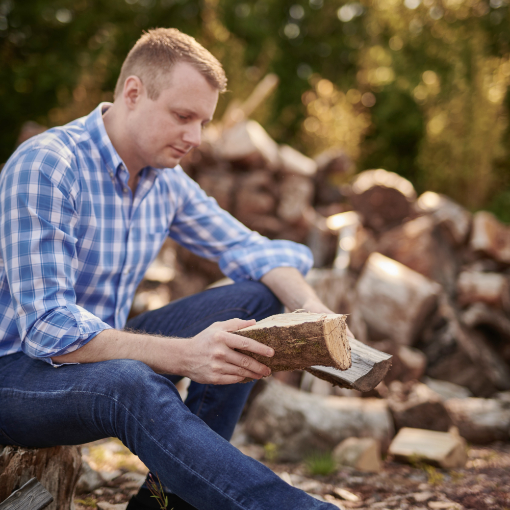 male wearing blue and white check shirt and jeans sitting on log holding pieces of chopped timber