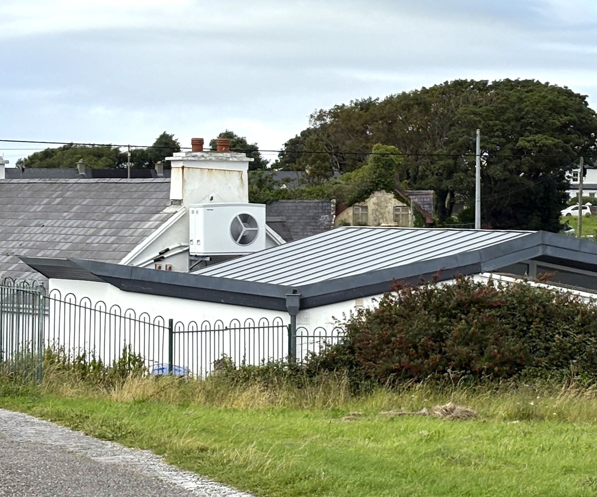 White air source heat pump mounted on an exterior wall of a white house with slate roof, visible behind a modern flat-roofed extension.