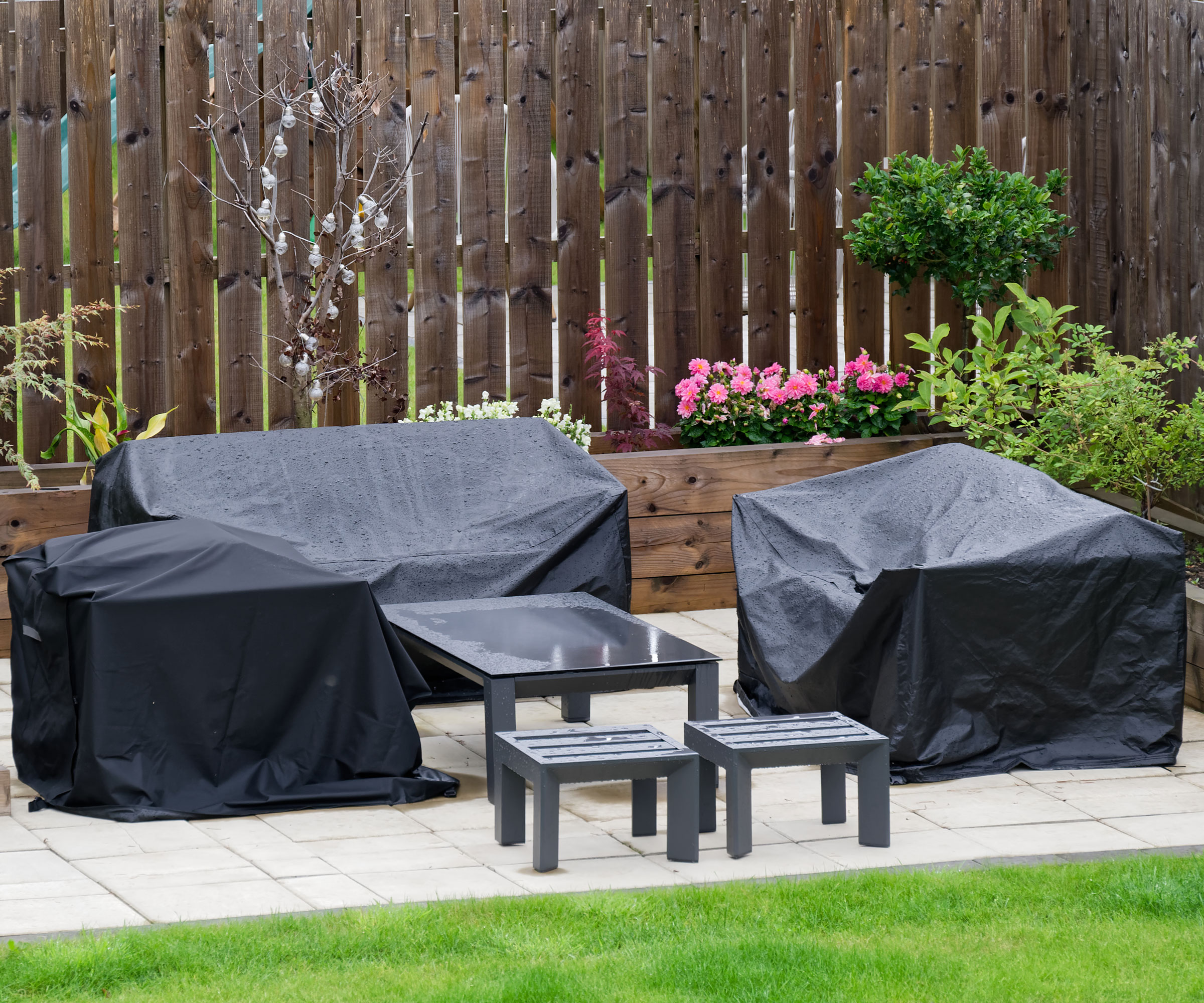 patio with garden furniture covered up for winter, with raised bed planted with flowers and shrubs, and a fence in the background plus lawn in the foreground