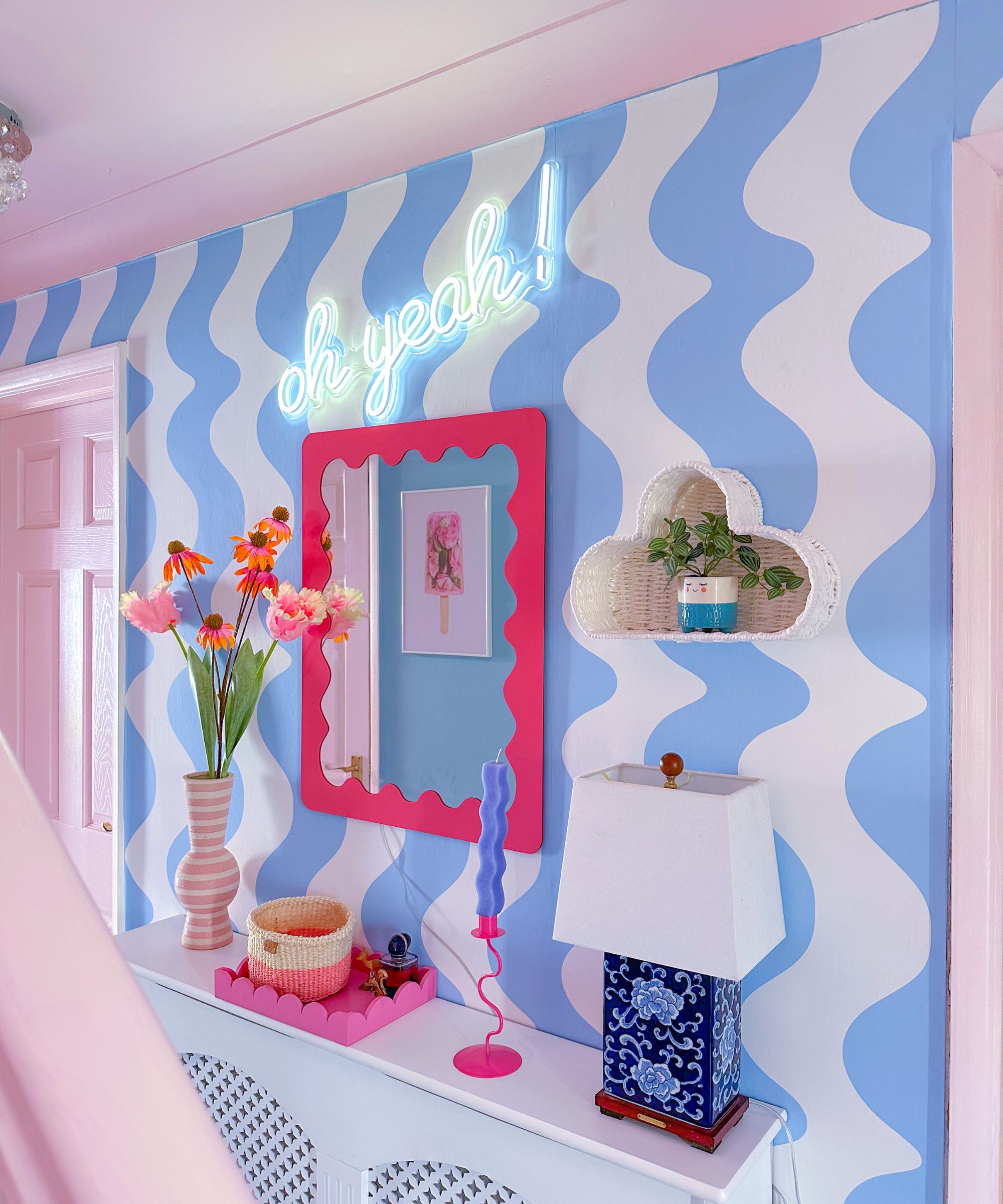 A hallway wall painted with bold blue and white wavy stripes. A hot pink mirror, neon sign saying &ldquo;oh yeah!&rdquo;, pink vase with flowers, and a quirky blue lamp add a playful retro feel.