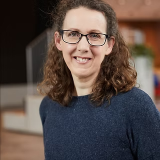 female with curly shoulder length hair wearing glasses and navy jumper
