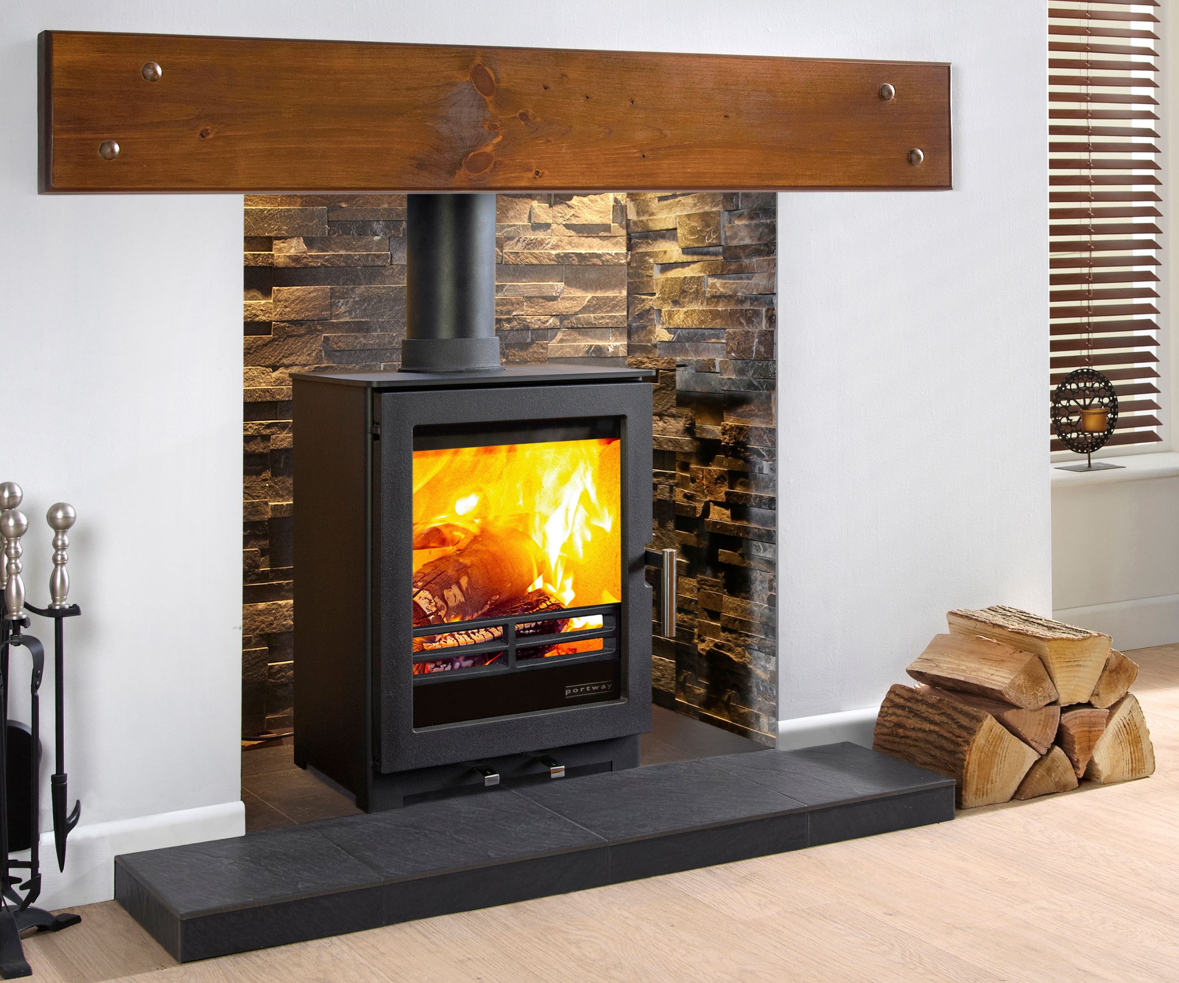 A black log burner with bright flames is framed by a stone-effect fireplace. A chunky wooden mantel stretches across the opening, with stacked firewood placed neatly to the side. The hearth is made of dark slate tiles.