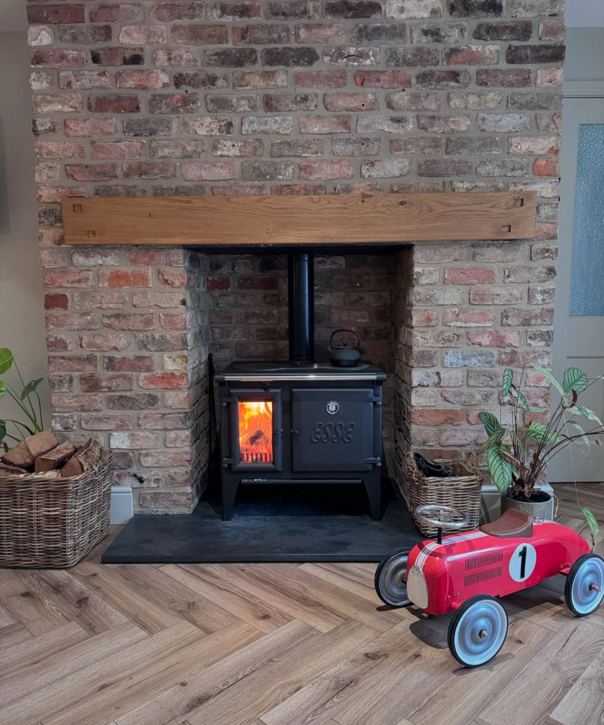 A rustic exposed brick fireplace with a black cast-iron log burner beneath a solid oak mantel beam. A wicker basket of logs sits nearby, and a child’s red vintage-style toy car rests on the wooden herringbone floor, adding a playful, homely touch.
