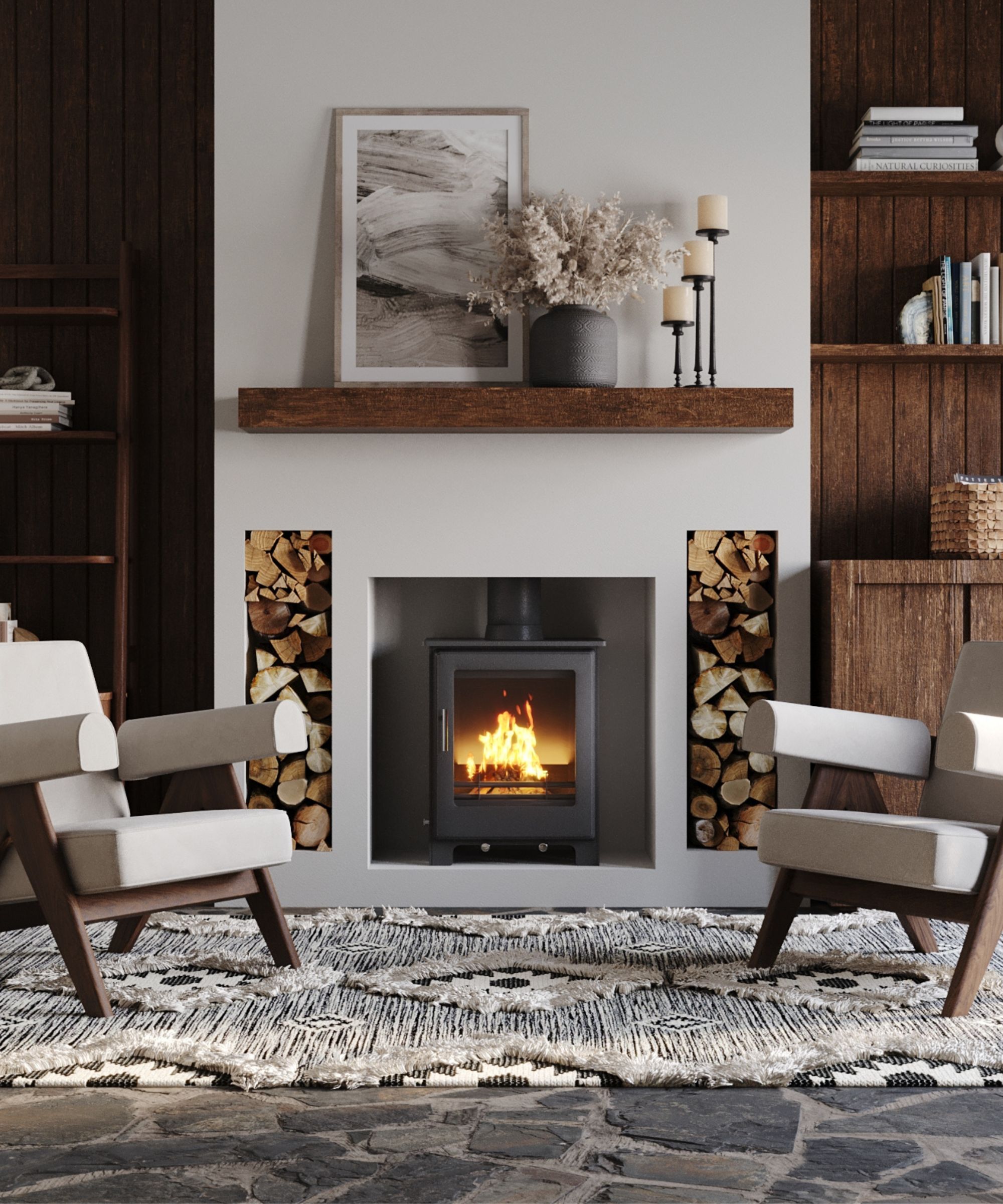 A modern living room with a sleek black log burner set into a minimalist white wall. The fireplace is framed by cubby holes filled with neatly stacked firewood. A chunky oak beam mantel above holds a monochrome abstract artwork and a grey vase of dried flowers, adding rustic charm