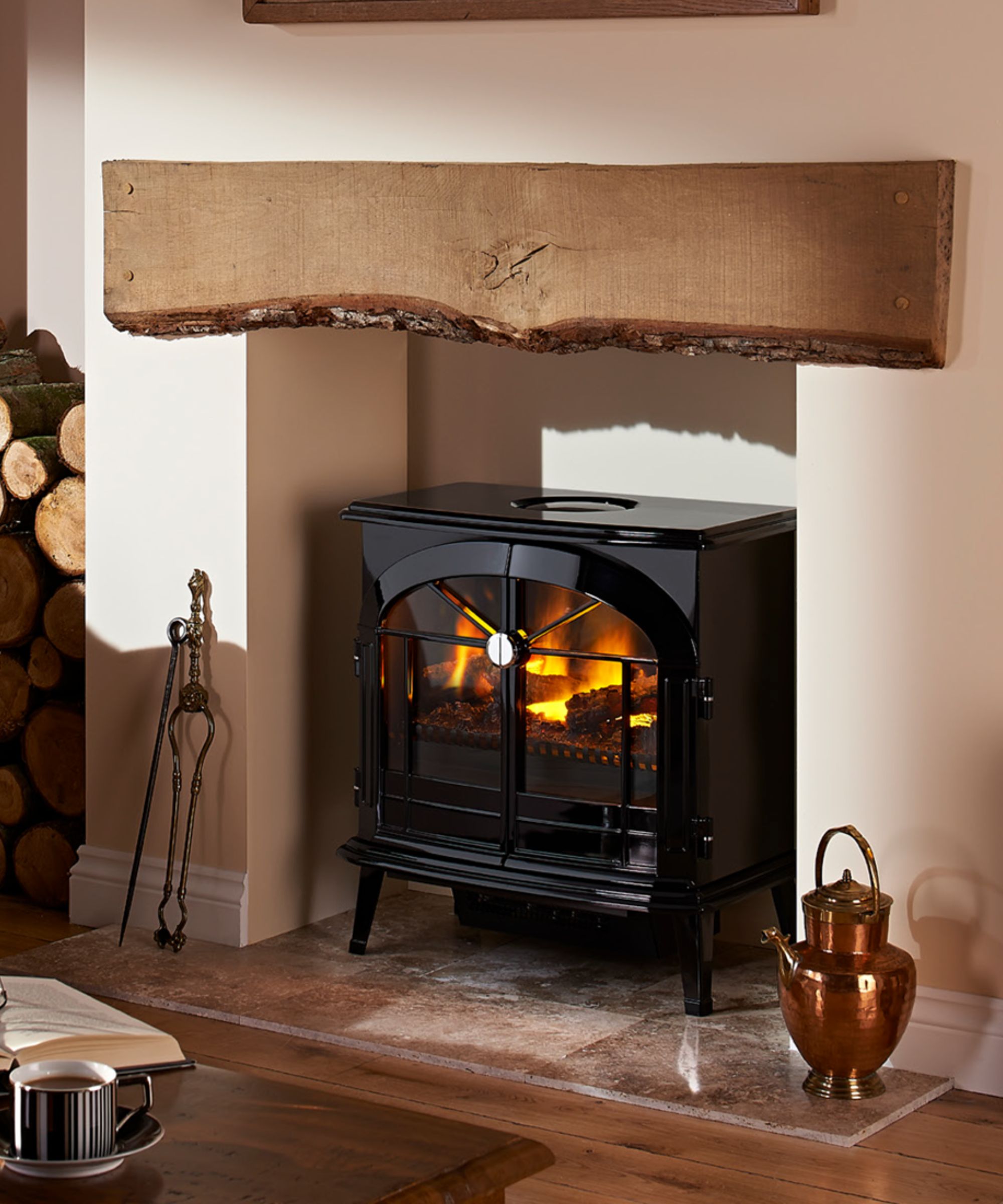 A traditional-style fireplace with a polished black cast-iron stove set on a stone hearth. Above it, a chunky oak beam mantel has been left with its raw bark edge intact, adding rustic character. A copper kettle sits beside the fire, while stacked logs and a wooden floor enhance the cosy cottage feel.