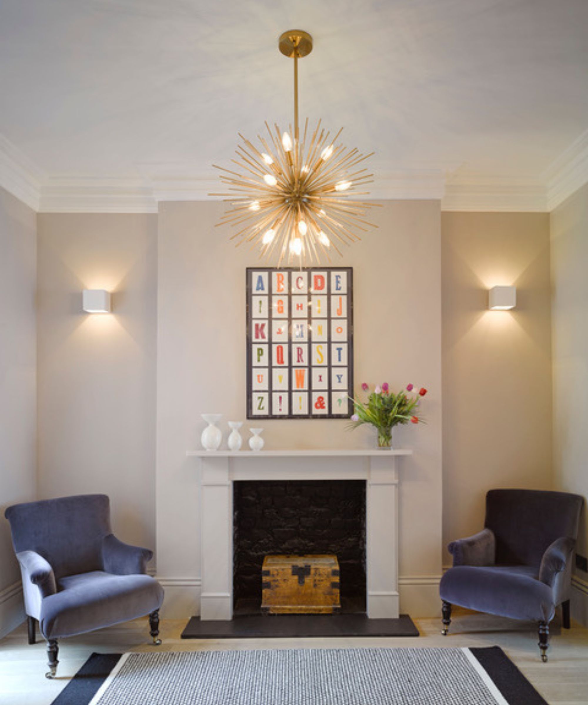 A light-filled living room with a bold and elegant design. Two deep blue velvet armchairs flank a white-painted fireplace, above which hangs a colourful alphabet artwork. A dramatic gold starburst chandelier takes centre stage