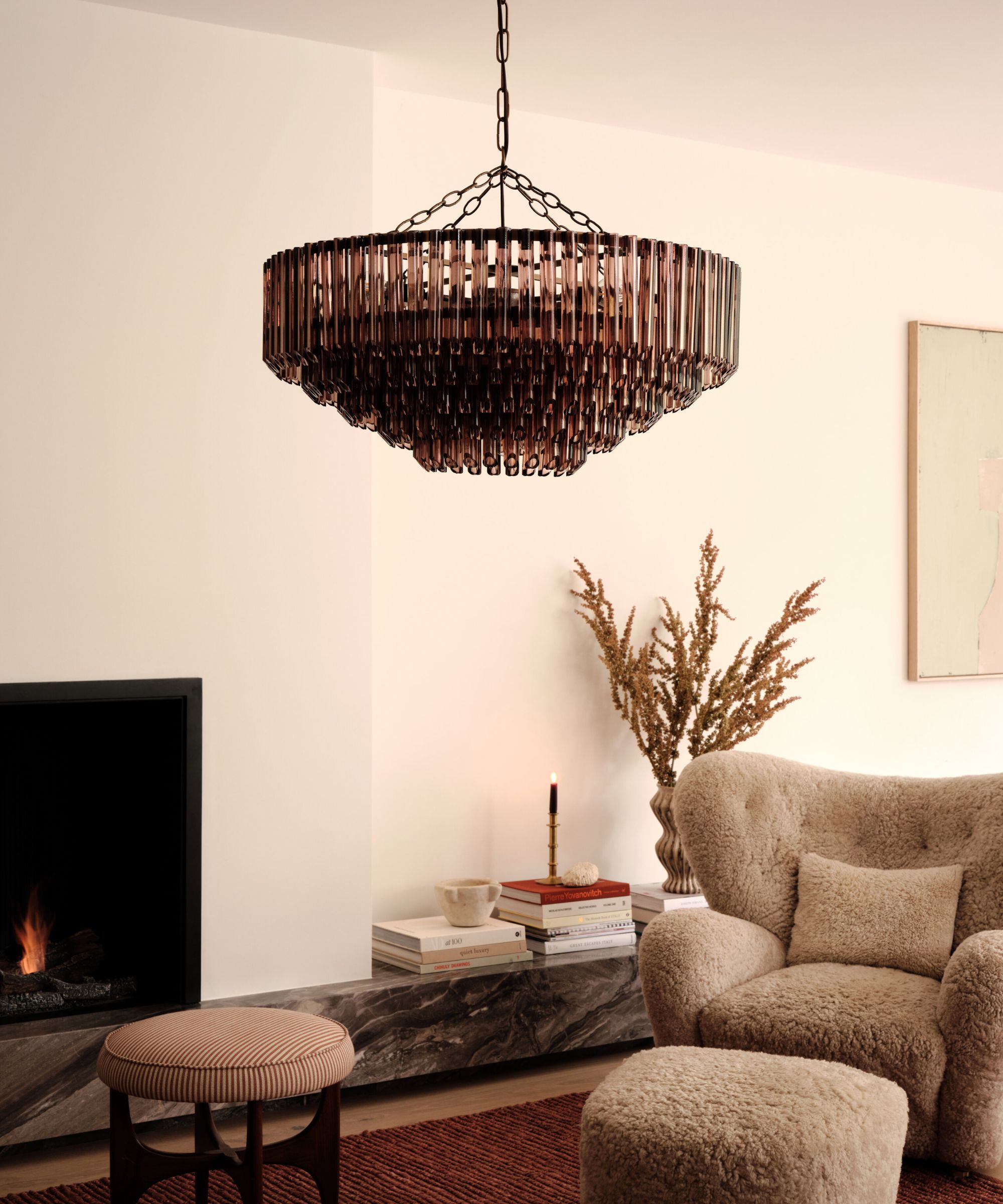 A warm, elegant living space featuring a plush beige armchair and matching footstool in a soft, textured fabric. Above hangs a striking dark glass chandelier with tiered details, suspended from a chain.