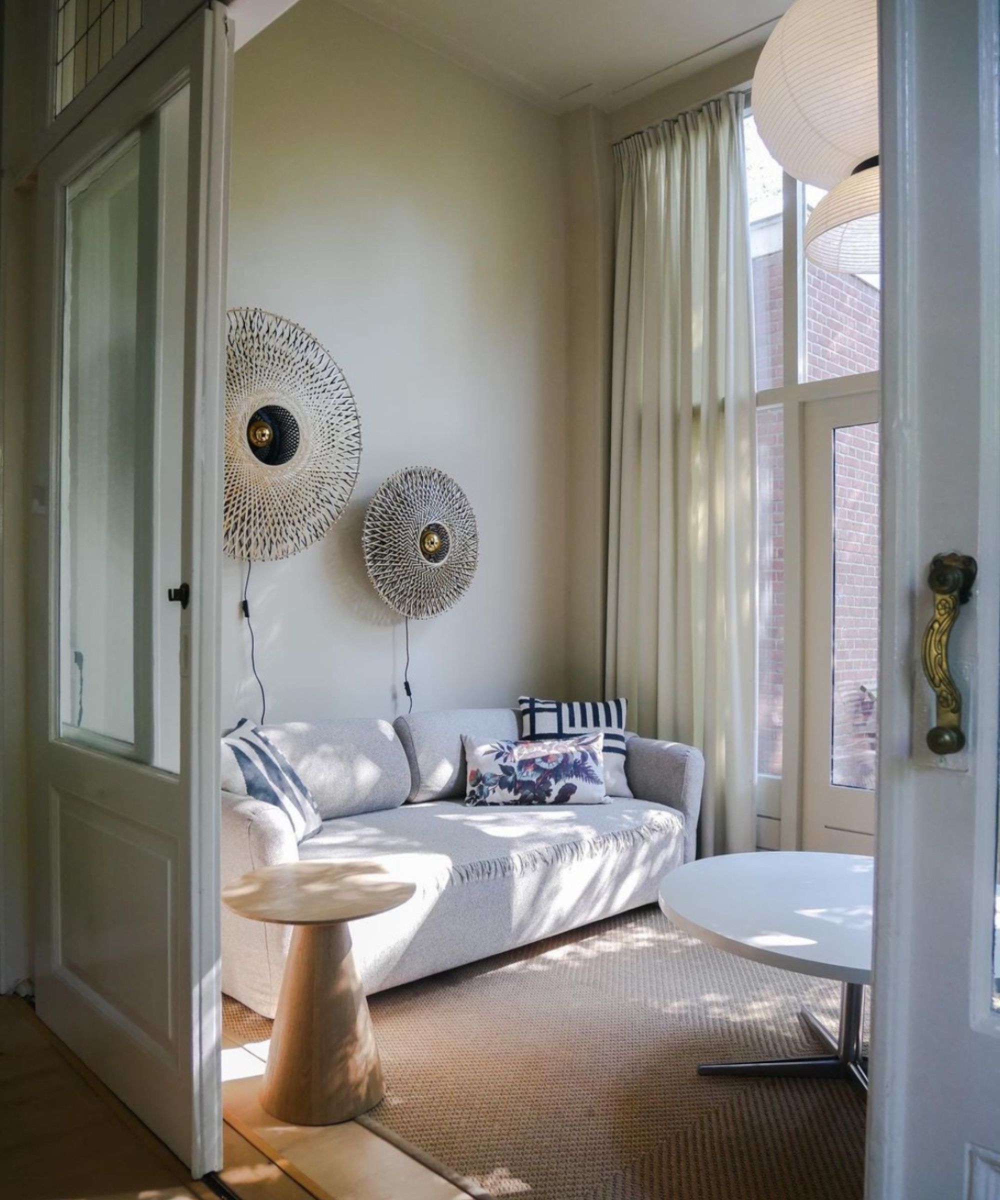 A bright, cosy sitting nook with a light grey sofa decorated with striped and floral cushions, placed against a cream wall. Two woven circular wall lights add texture above the sofa, while sheer floor-to-ceiling curtains frame tall glass doors.
