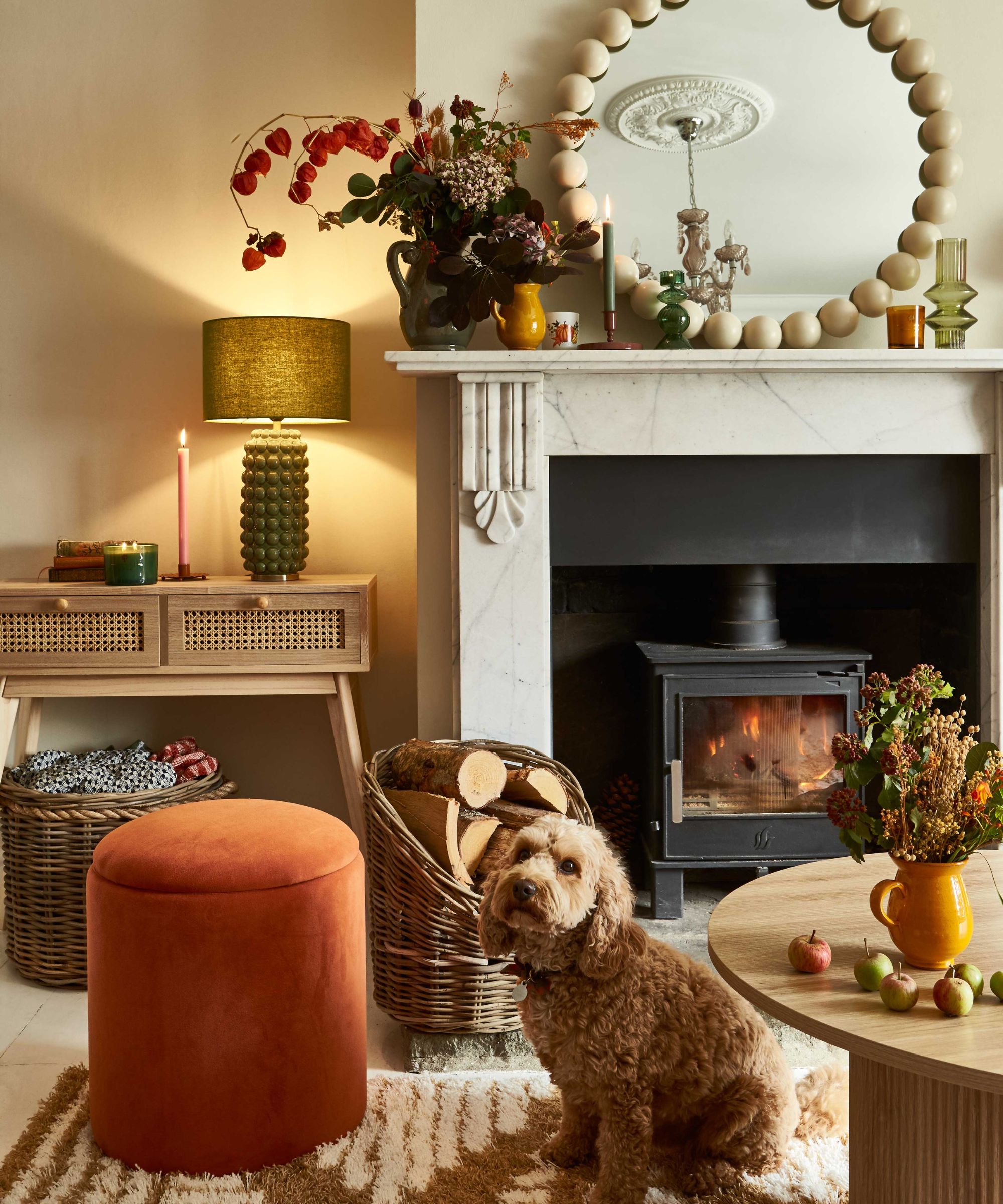 A cosy living room corner styled with warm, autumnal tones. A fluffy brown dog sits on a patterned rug in front of a wood-burning stove set into a white marble fireplace. On the mantelpiece are a round mirror with a bold beaded frame, candles, and vases of flowers.