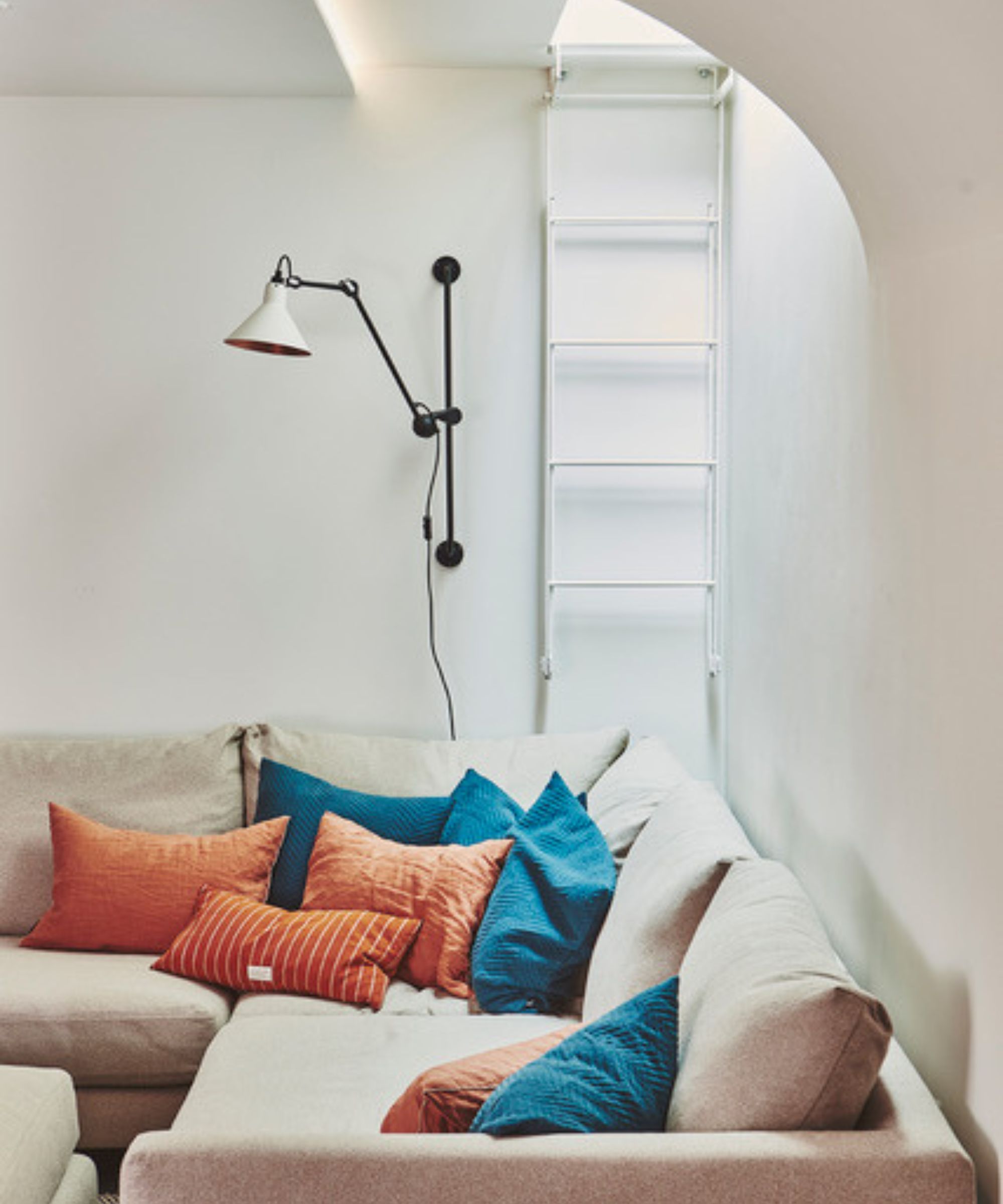 A compact living room corner featuring a neutral L-shaped sofa piled with bold orange and teal cushions. A black adjustable wall-mounted lamp with a conical shade provides task lighting above. To the side, a slim white ladder leads up to a ceiling hatch, adding a quirky, functional detail to the space.