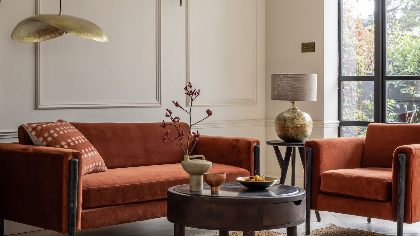A stylish modern living room with warm, earthy tones. It features a burnt orange velvet sofa and matching armchair with black wooden frames. A round dark wood coffee table sits in the centre, decorated with ceramic vases and a bowl of fruit. To the side, a brass-textured table lamp with a neutral shade rests on a small side table.