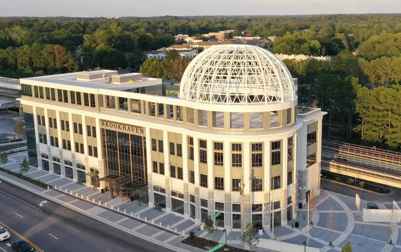 A large, newly constructed building looms above the treeline, with an open roof space and cars around.