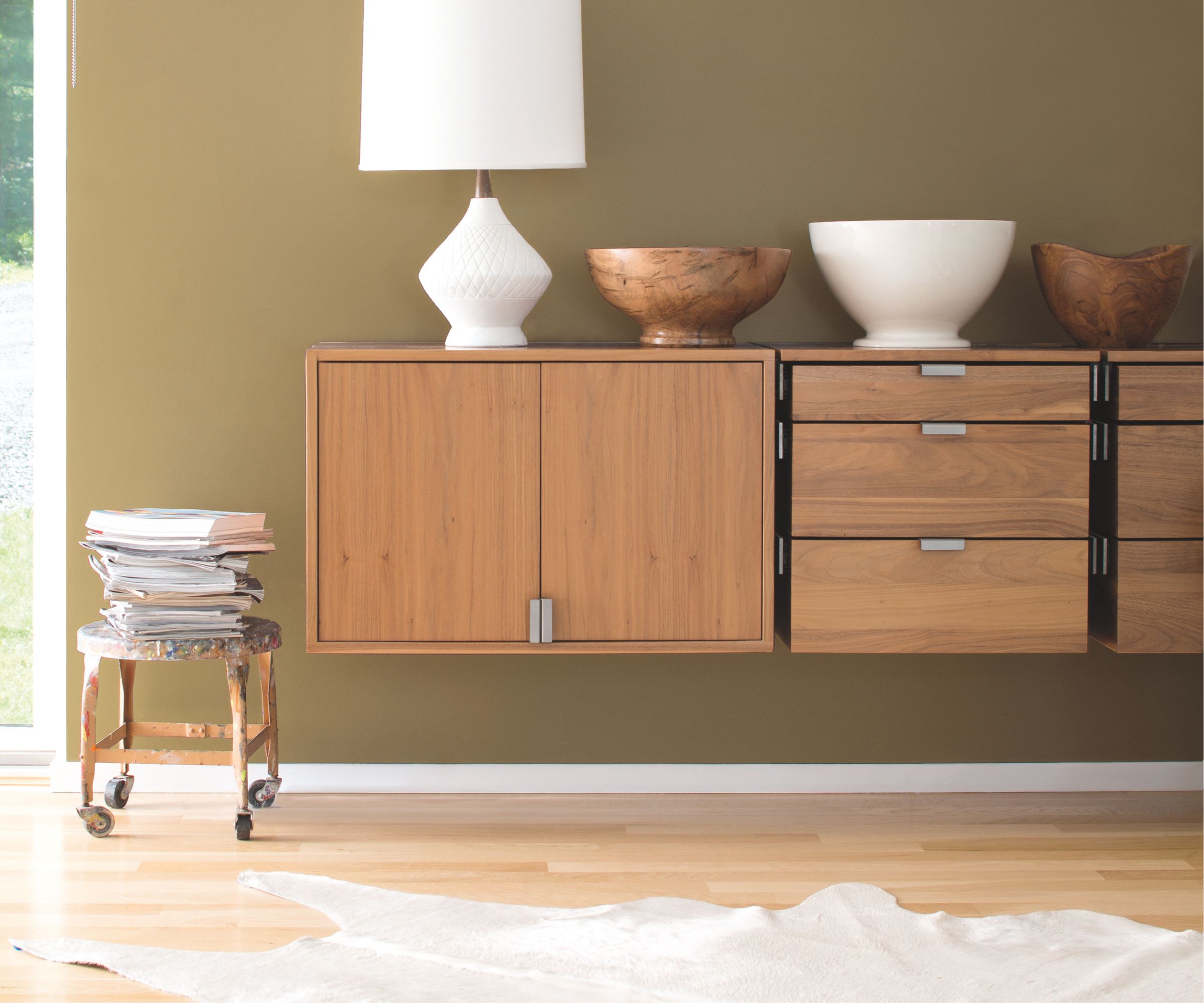 A modern interior with floating wooden cabinets against an olive-green wall. The units are topped with decorative bowls and a white lamp, while a stack of magazines sits on a rustic stool nearby.