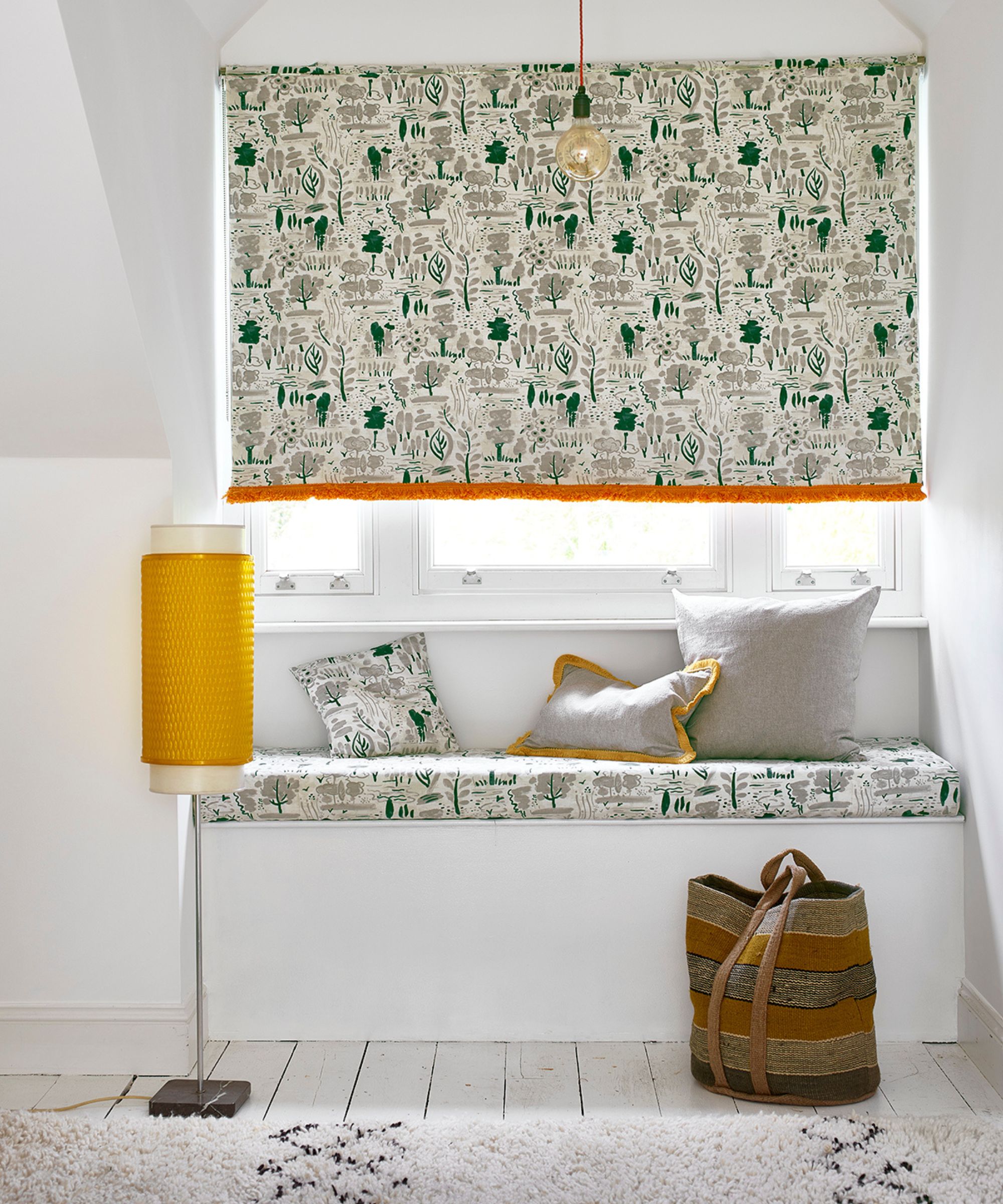 A bright, cosy window nook features a seat upholstered in patterned fabric with green and beige woodland motifs, paired with matching cushions. A soft grey pillow adds contrast, while a mustard yellow floor lamp and striped woven bag bring warmth to the space.