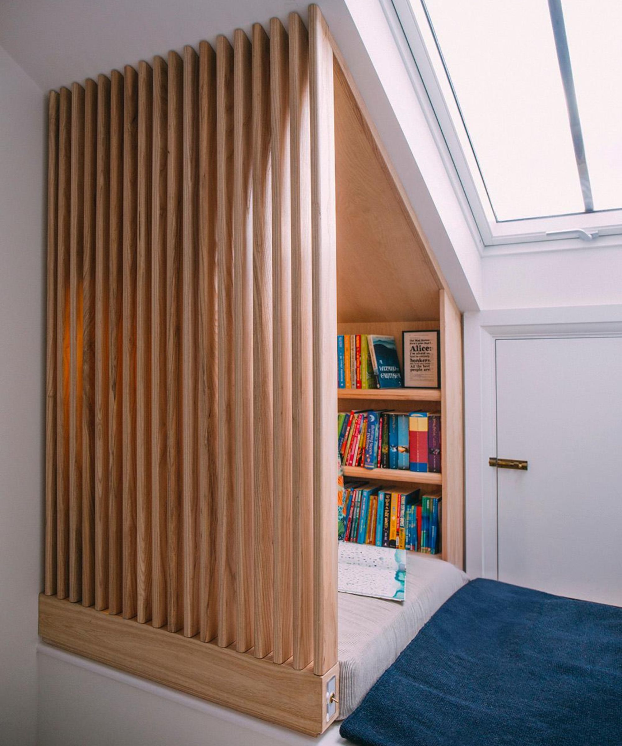 A small reading nook under a sloped ceiling features a built-in bed framed with vertical wooden slats. Natural light pours in through a skylight above, illuminating shelves filled with colourful books.