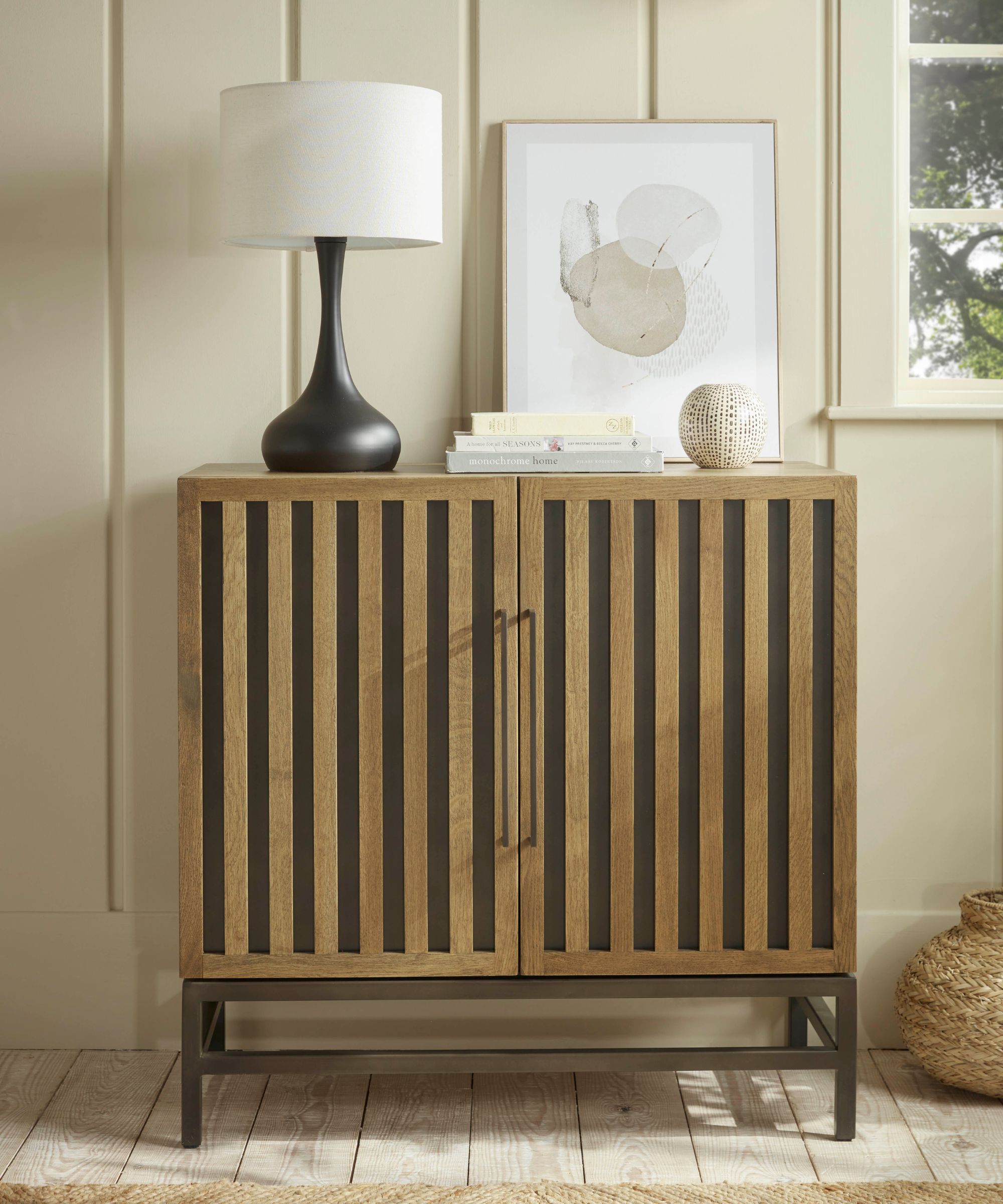 Compact wooden hallway cabinet with vertical slatted doors and slim black metal legs. Styled with a sculptural black lamp, abstract artwork, and neutral decor, it creates a modern, welcoming corner.