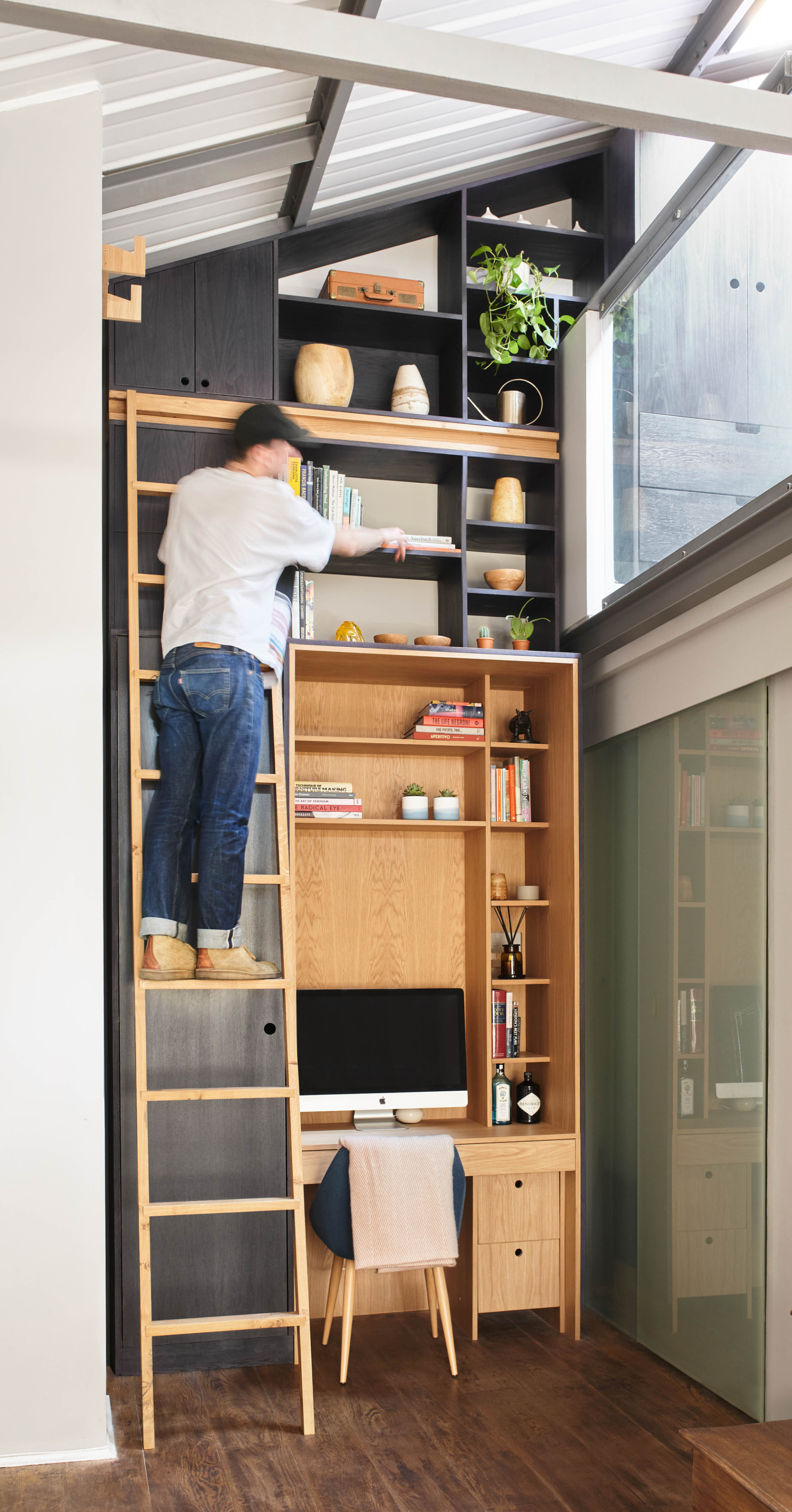 A tall wooden shelving unit with a built-in desk fills a modern room with a sloping ceiling. A man stands on a ladder, reaching for books on the upper shelves. The unit features open compartments, some filled with books, plants, and ceramics, and a computer sits on the desk below.