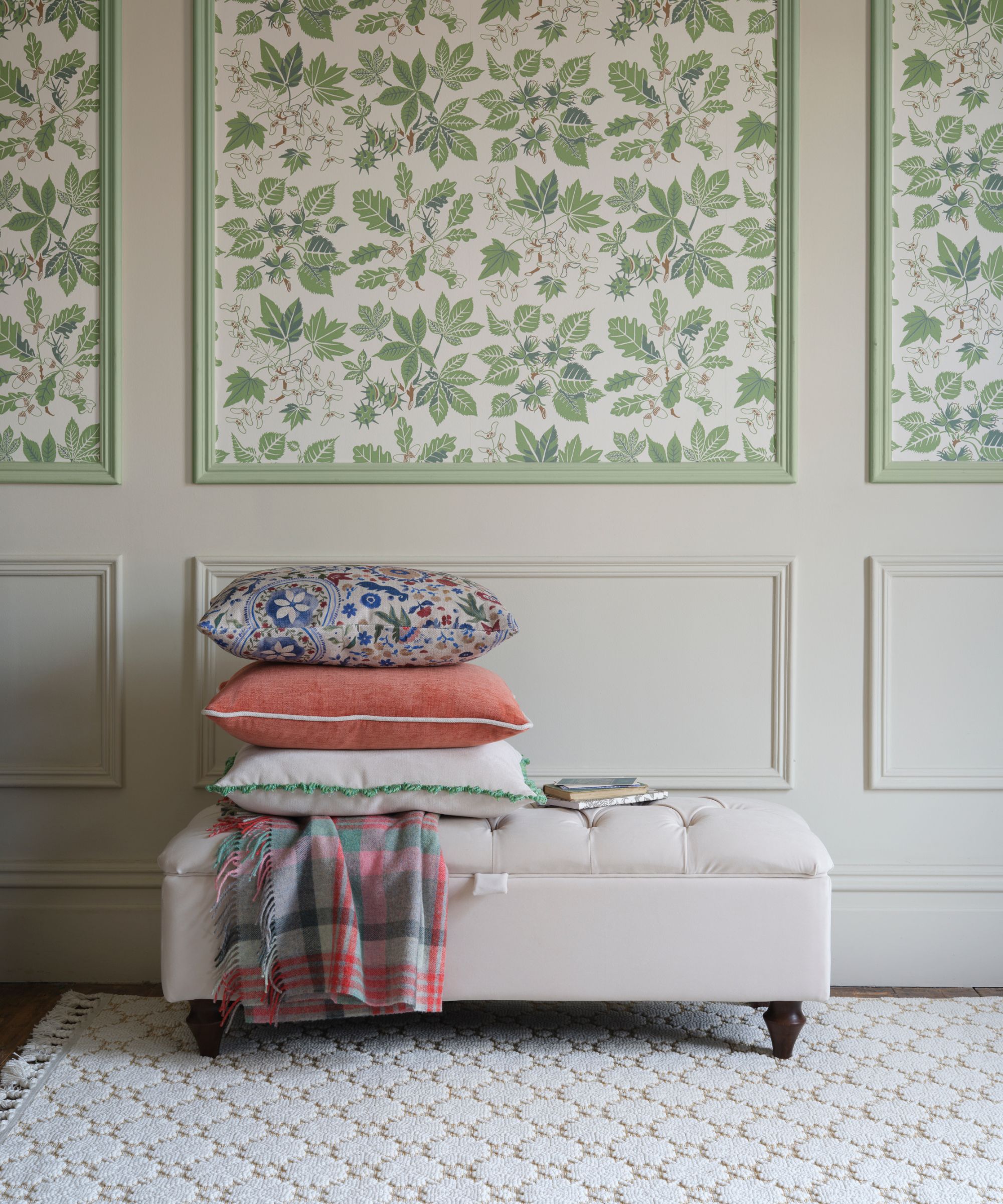 A traditional hallway corner with pale green panelled walls and matching floral wallpaper panels. A cushioned bench topped with stacked pillows and a folded blanket adds a cosy, classic touch.