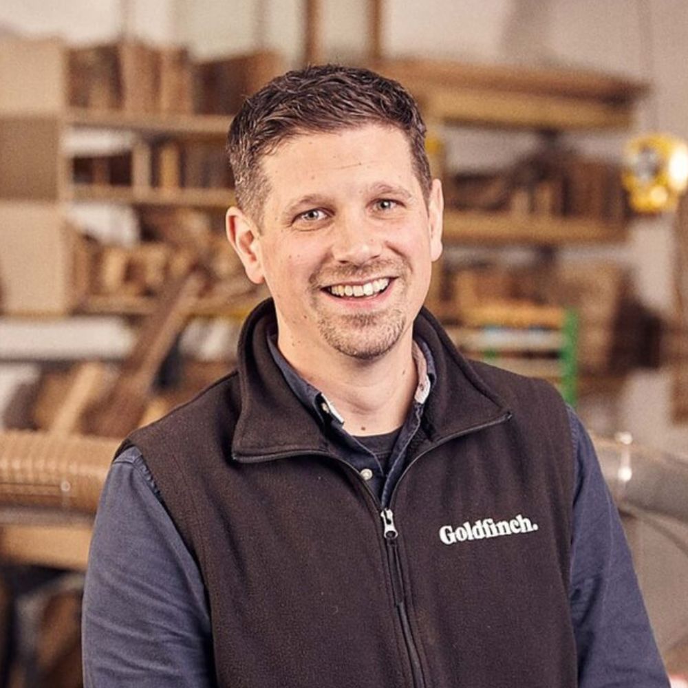 A smiling man stands in a workshop surrounded by wooden materials and tools. He’s wearing a dark fleece vest with the word “Goldfinch” embroidered on it over a collared shirt.
