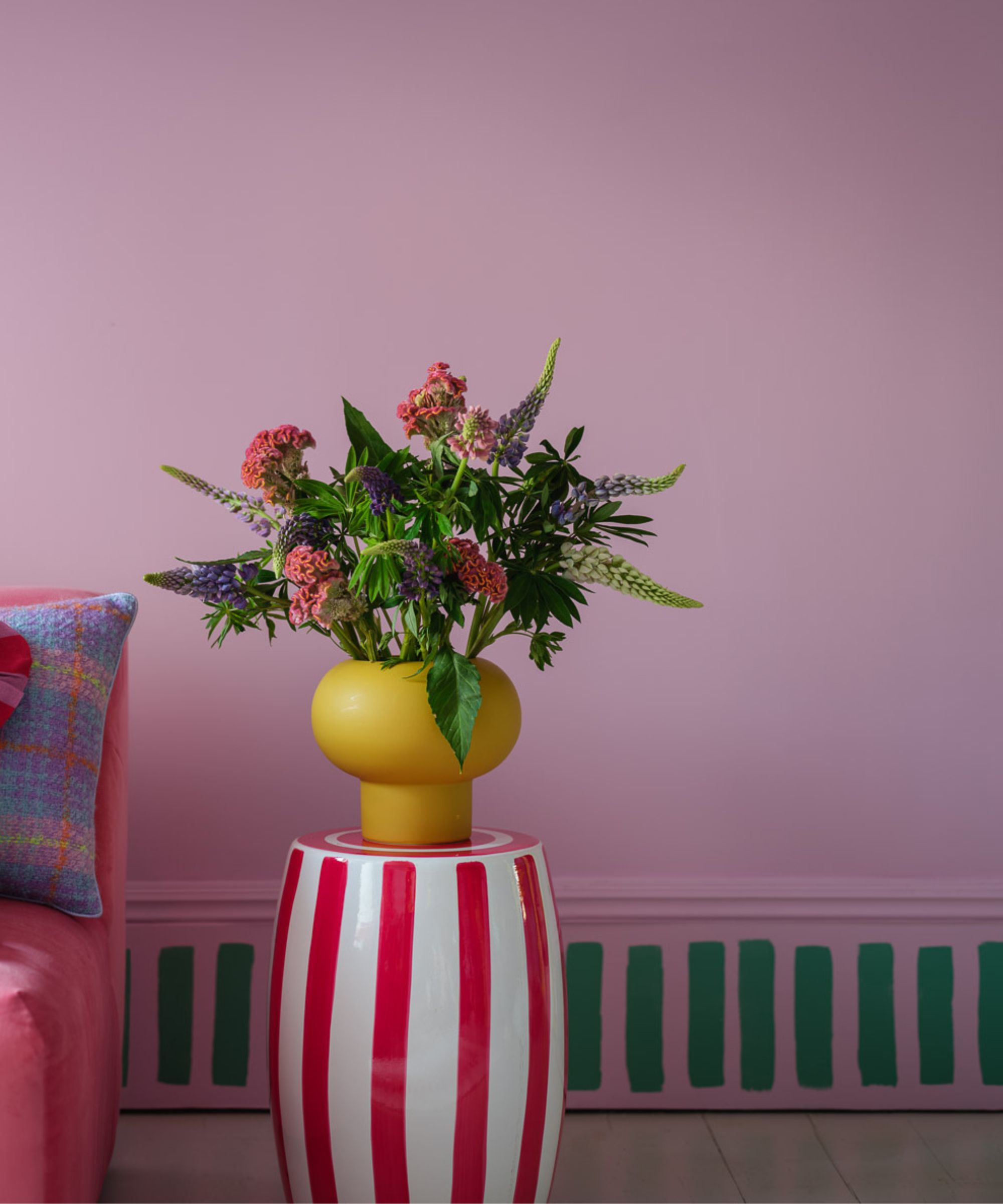 A playful, colourful interior featuring a pink wall with a vibrant striped skirting board painted in green and pink. A bold red-and-white striped side table holds a bright yellow vase filled with colourful flowers.