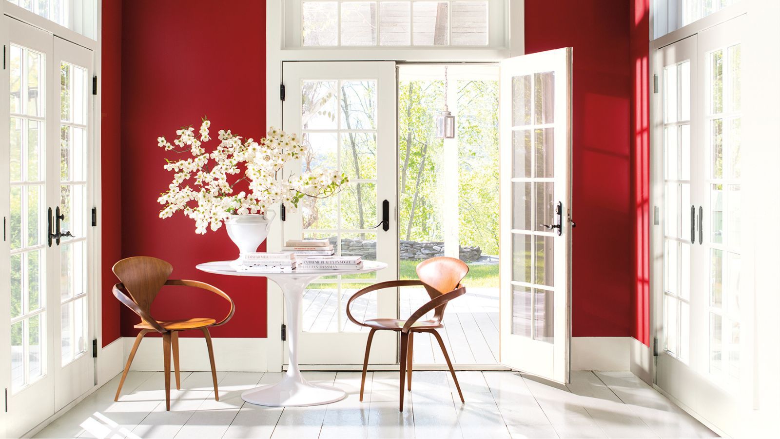 A bright sunroom with bold red walls and white French doors. Mid-century wooden chairs and a round white table topped with blooming branches create a striking yet inviting modern contrast.