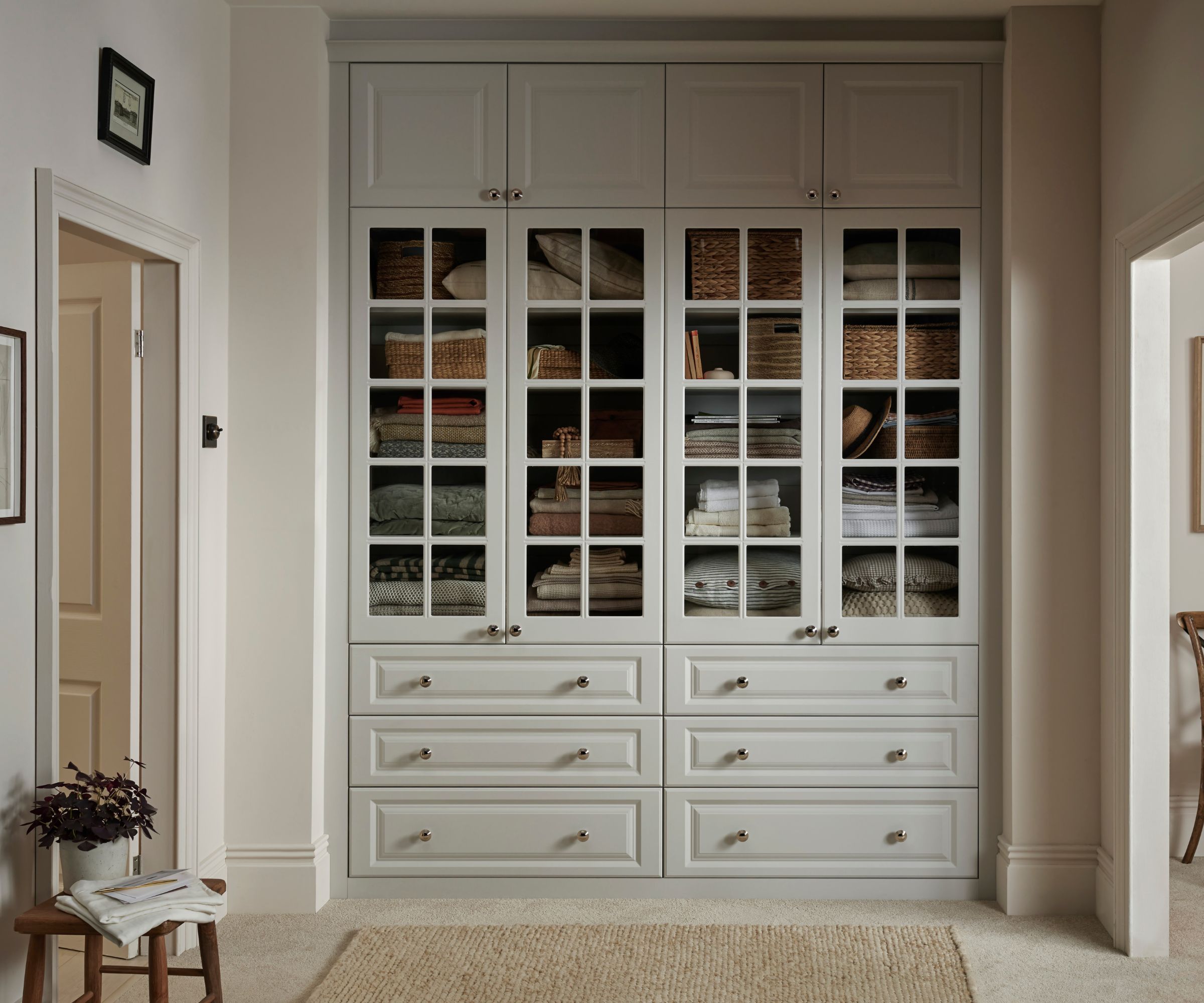 Elegant built-in hallway storage with floor-to-ceiling cabinetry in soft grey. Glass-fronted doors reveal neatly stacked linens and woven baskets, while lower drawers and upper cupboards keep the space organised and serene.