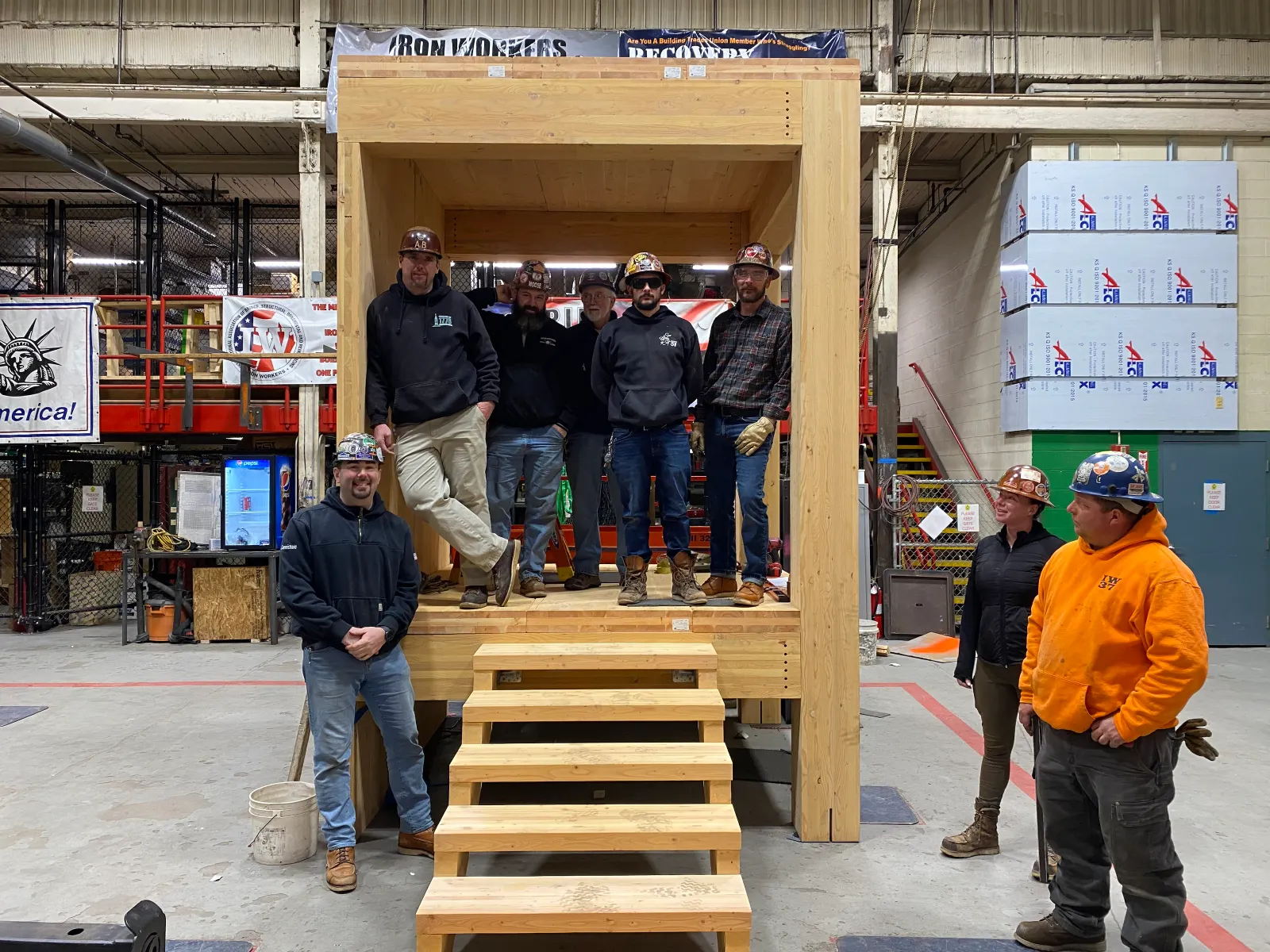 A group of people in jeans, hoodies and other work and safety gear stand within a small, wood enclosure.