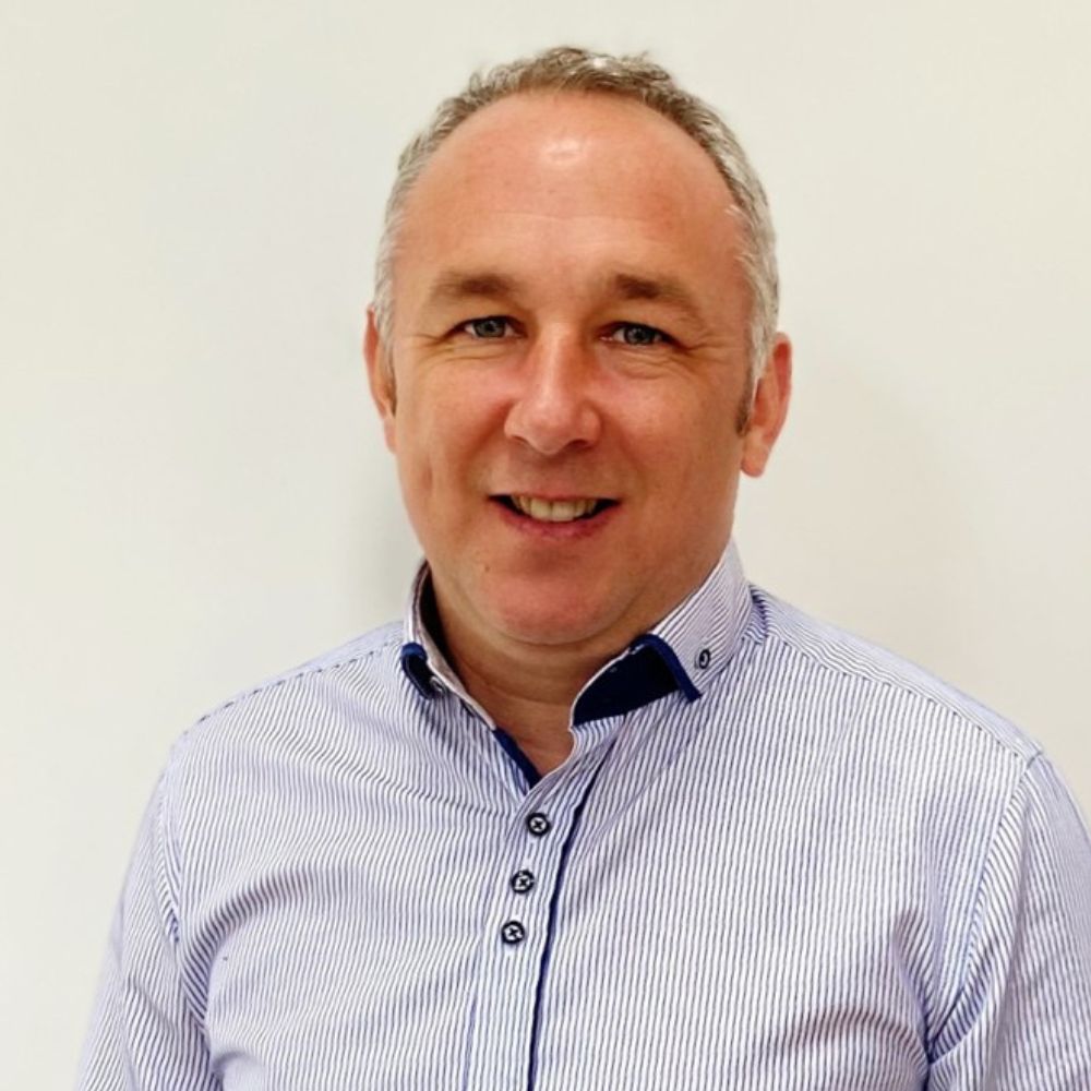 A smiling man wearing a light blue striped shirt stands against a plain white background.