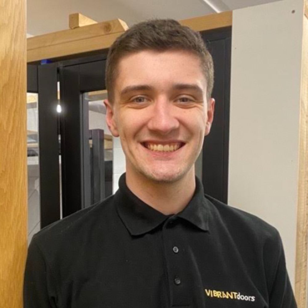 A man in a black polo shirt with a company logo smiles while standing indoors beside wooden door frames.