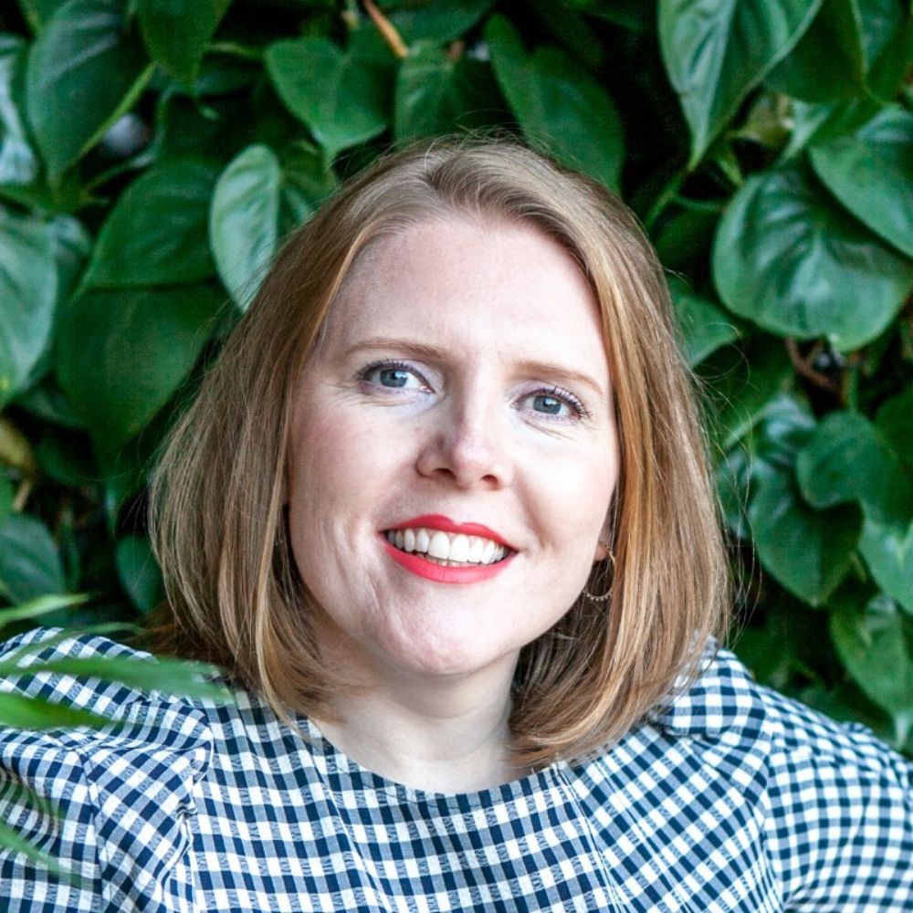 A woman with short hair and a checked top smiles while standing in front of green foliage.